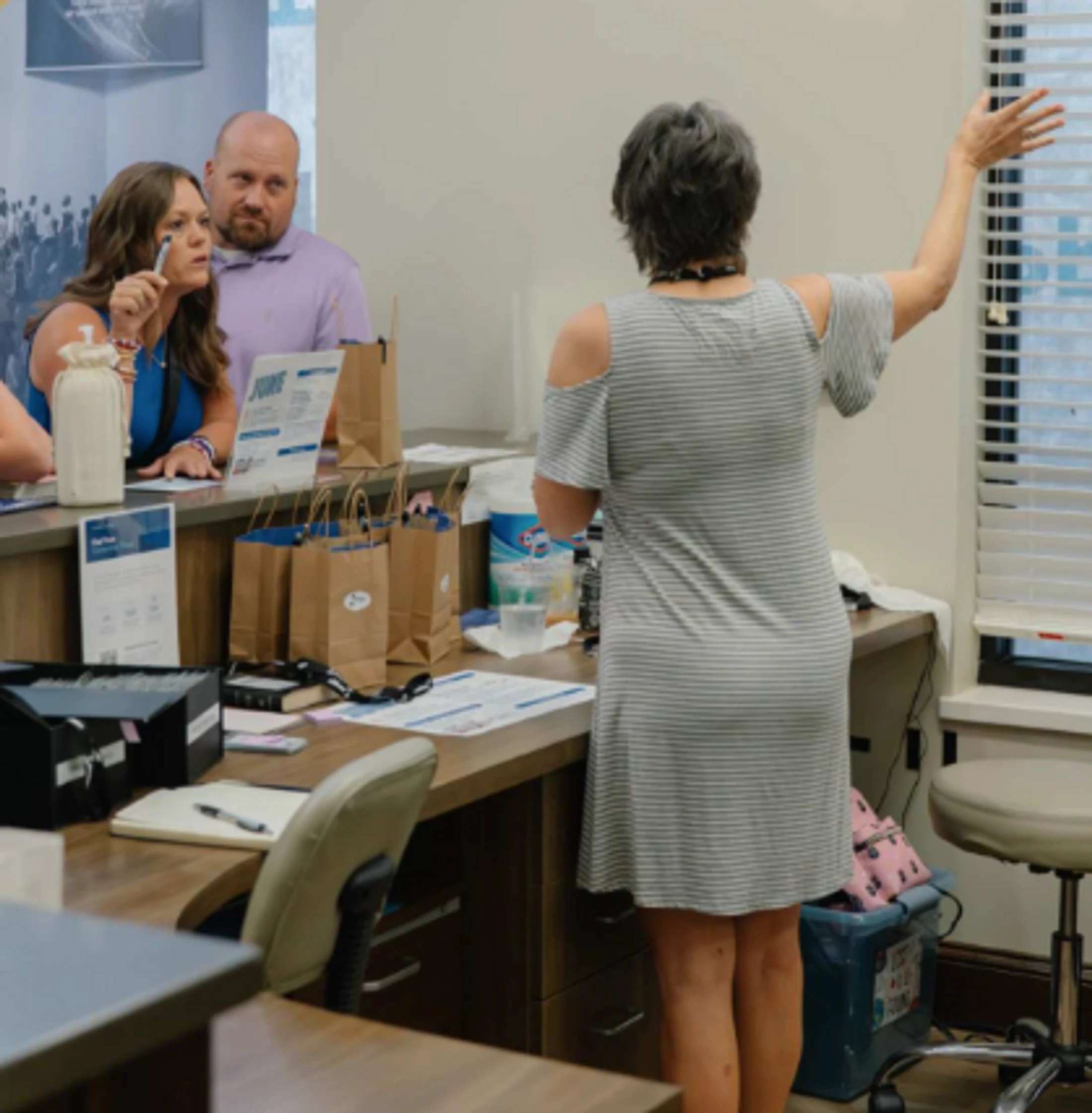 A woman in a striped dress gestures toward a window while talking to a man and another woman who are listening attentively from behind an office counter.