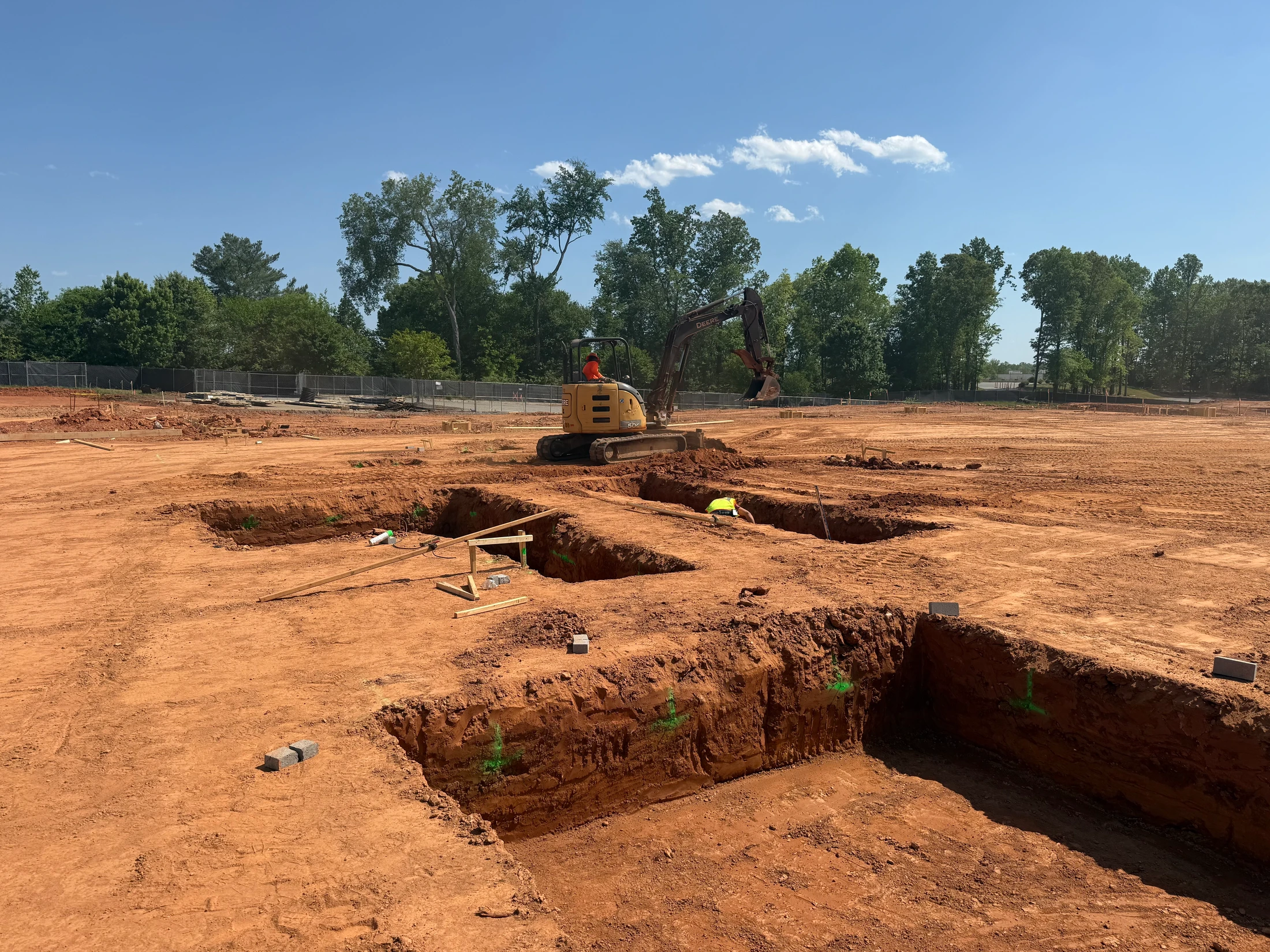A large construction site with trenches dug into red earth for a building's foundation. An excavator sits in the background and a worker is in a trench.