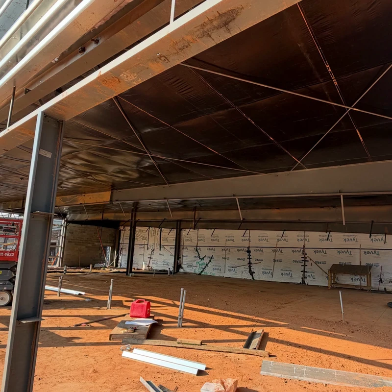 A wide shot of a building's interior during construction, with exposed steel beams, black ceiling insulation, a dirt floor, and walls covered in Tyvek wrap.