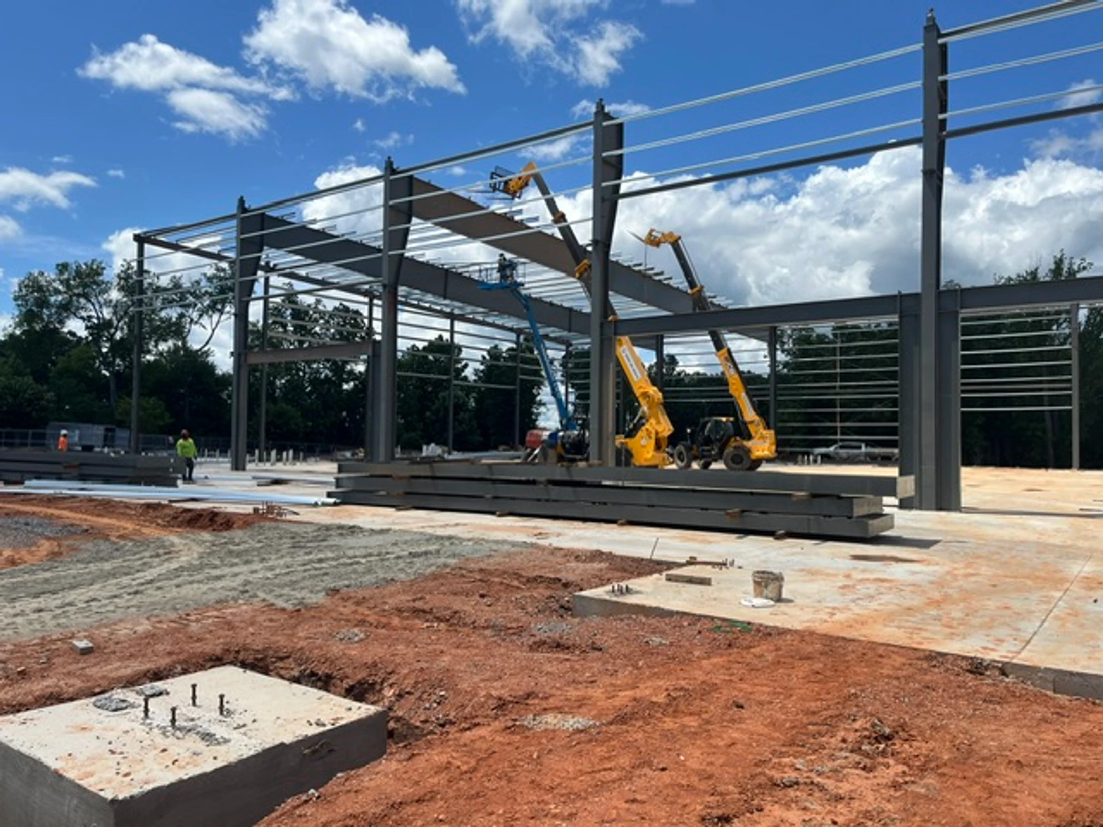 A construction site with the steel frame of a large building being erected. Yellow telehandlers and a blue lift are visible on the concrete foundation.