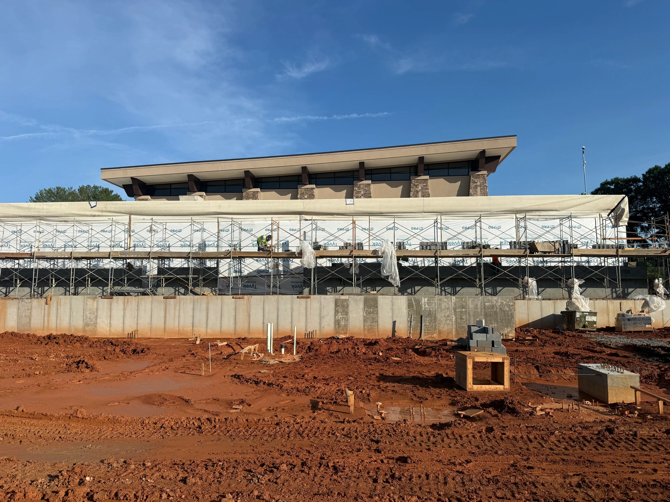 A modern building under construction with scaffolding against its foundation. A worker is on the scaffolding, and the foreground is muddy red earth.