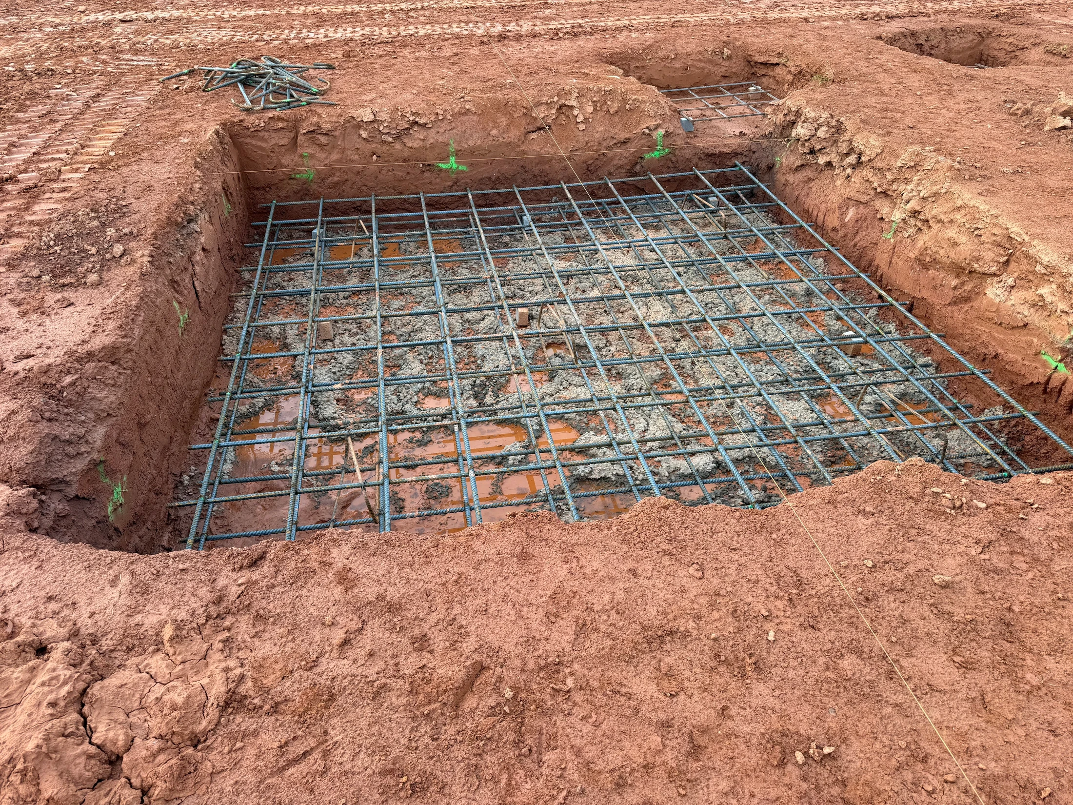 A square pit dug into red clay soil at a construction site, with a steel rebar grid laid inside, ready for a concrete pour.