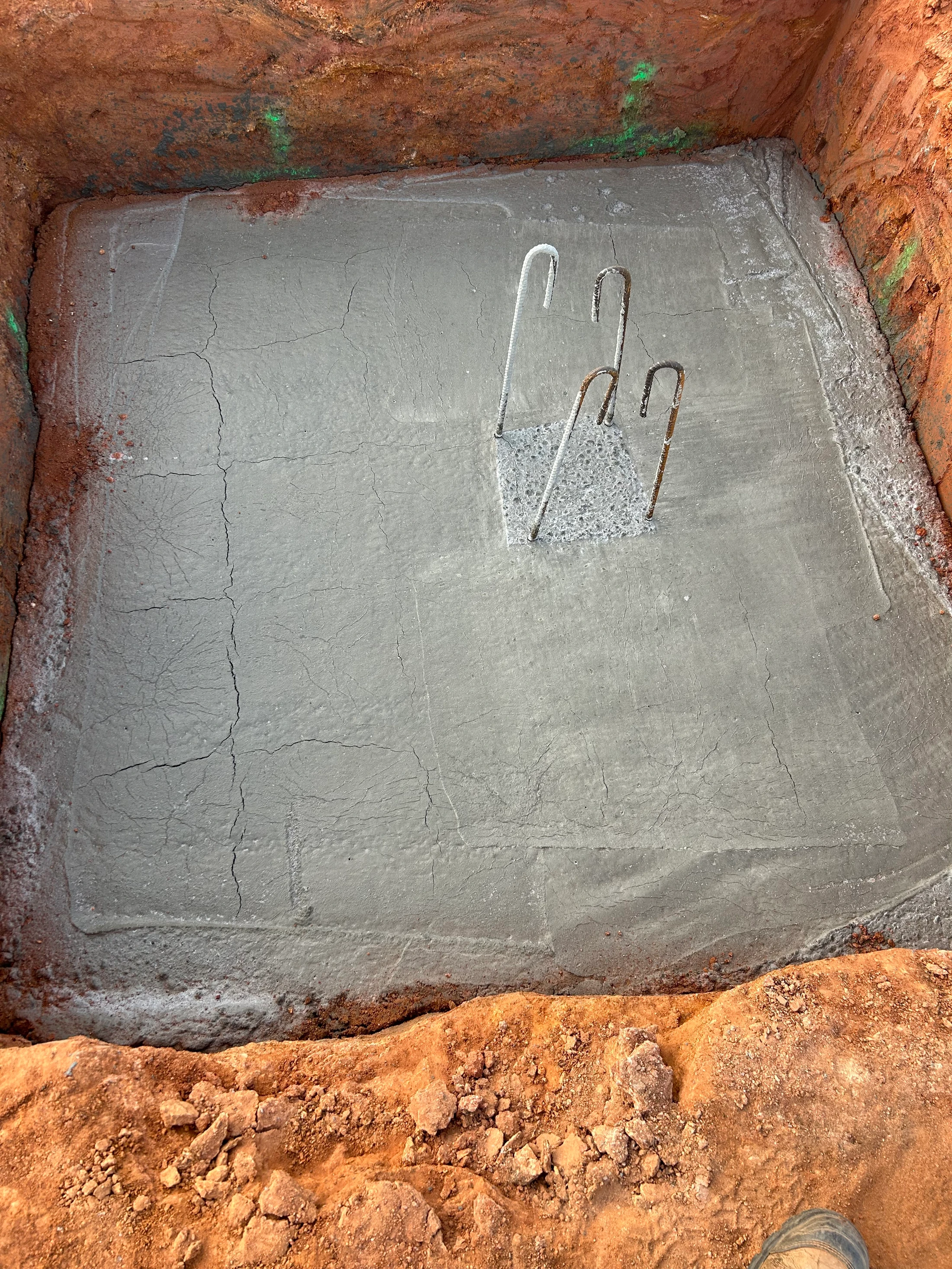A square construction pit dug into red earth, with a freshly poured concrete base that has surface cracks. Steel rebar hooks emerge from the concrete.