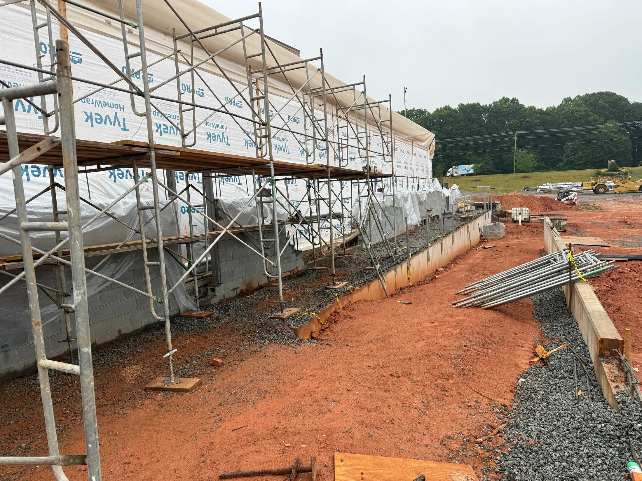 A long view of a building under construction with metal scaffolding erected along a concrete block wall. The ground is red dirt and gravel.
