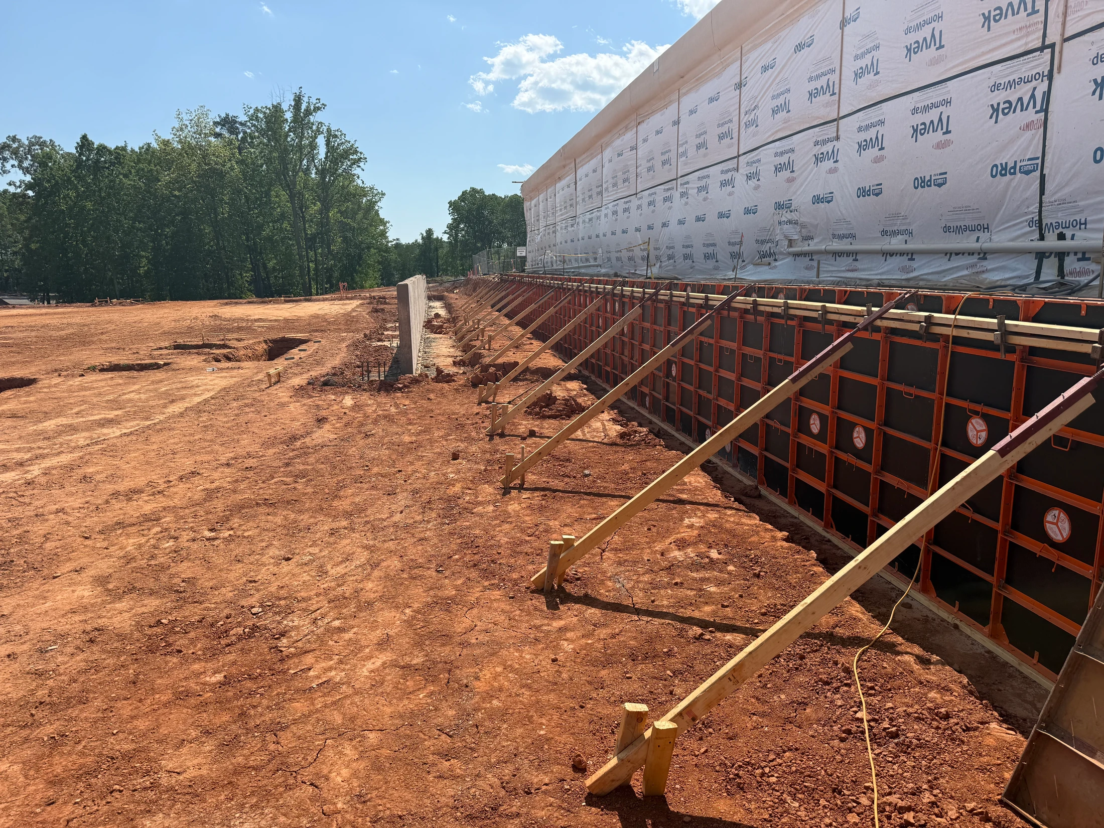 A construction site with red dirt ground. A long wall is being built, with one section showing concrete forms and another covered in white house wrap.