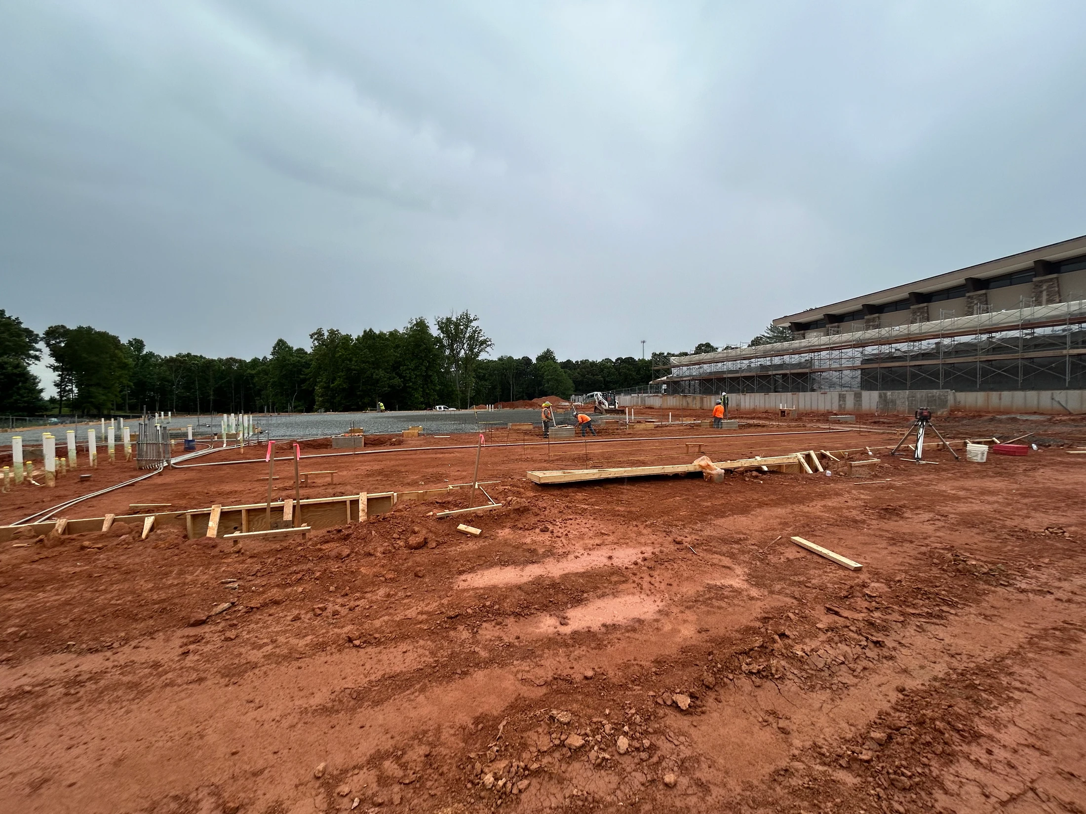 A wide view of a construction site with red dirt. Workers in orange vests prepare the foundation for a new building next to a larger structure with scaffolding.