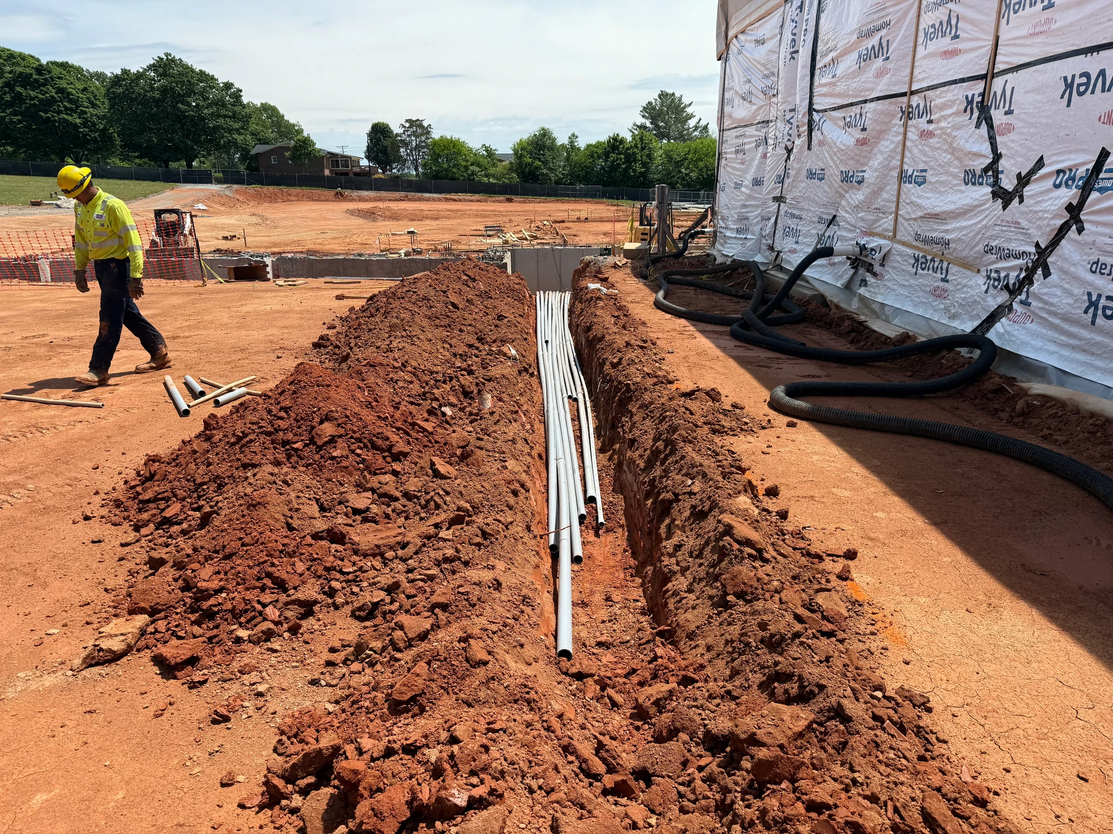 A construction worker walks past a freshly dug trench holding grey electrical conduit pipes on a red clay construction site next to a new building.