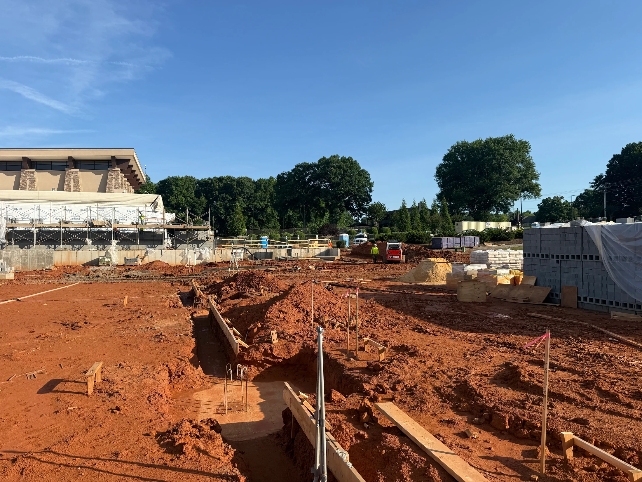 A large construction site with red clay soil, where the foundation of a new building is being laid next to an existing structure with scaffolding.