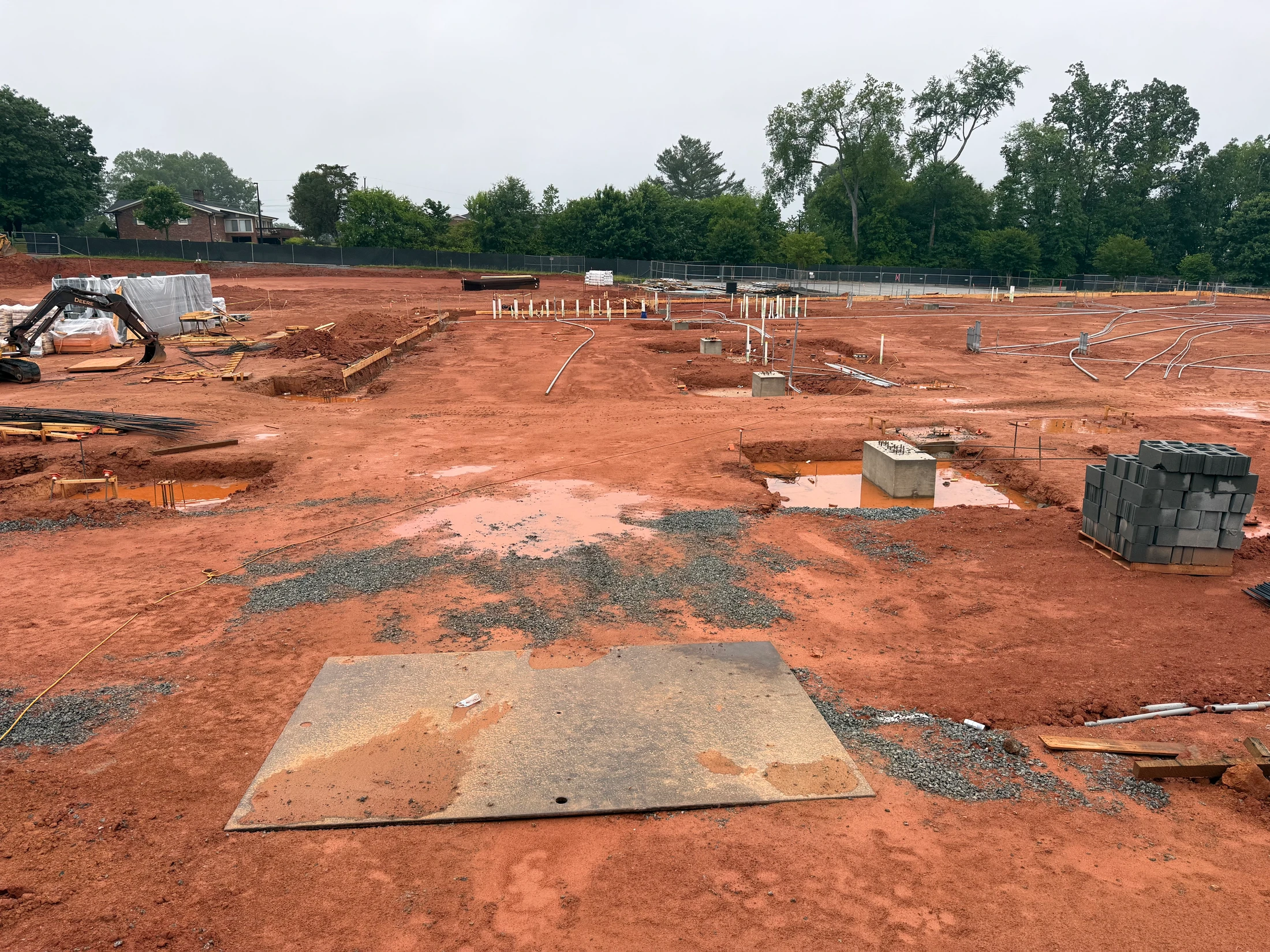A large construction site with wet, red clay ground. Concrete footings, plumbing pipes, and building materials are laid out for a new foundation.