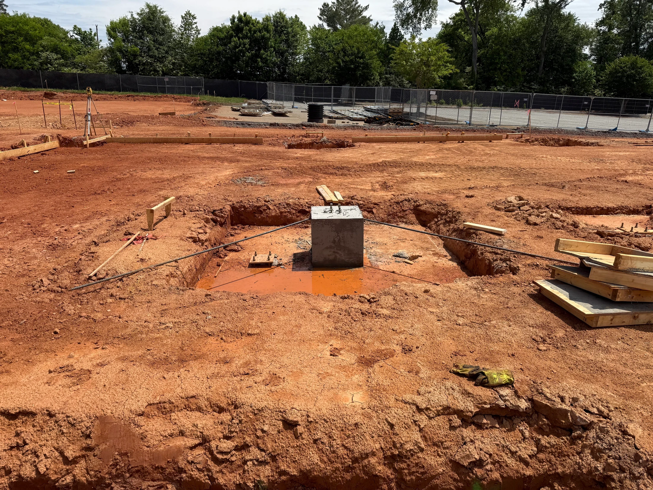 A construction site with excavated red clay. In the center, a concrete pier footing with anchor bolts sits in a muddy pit, braced by steel cables.