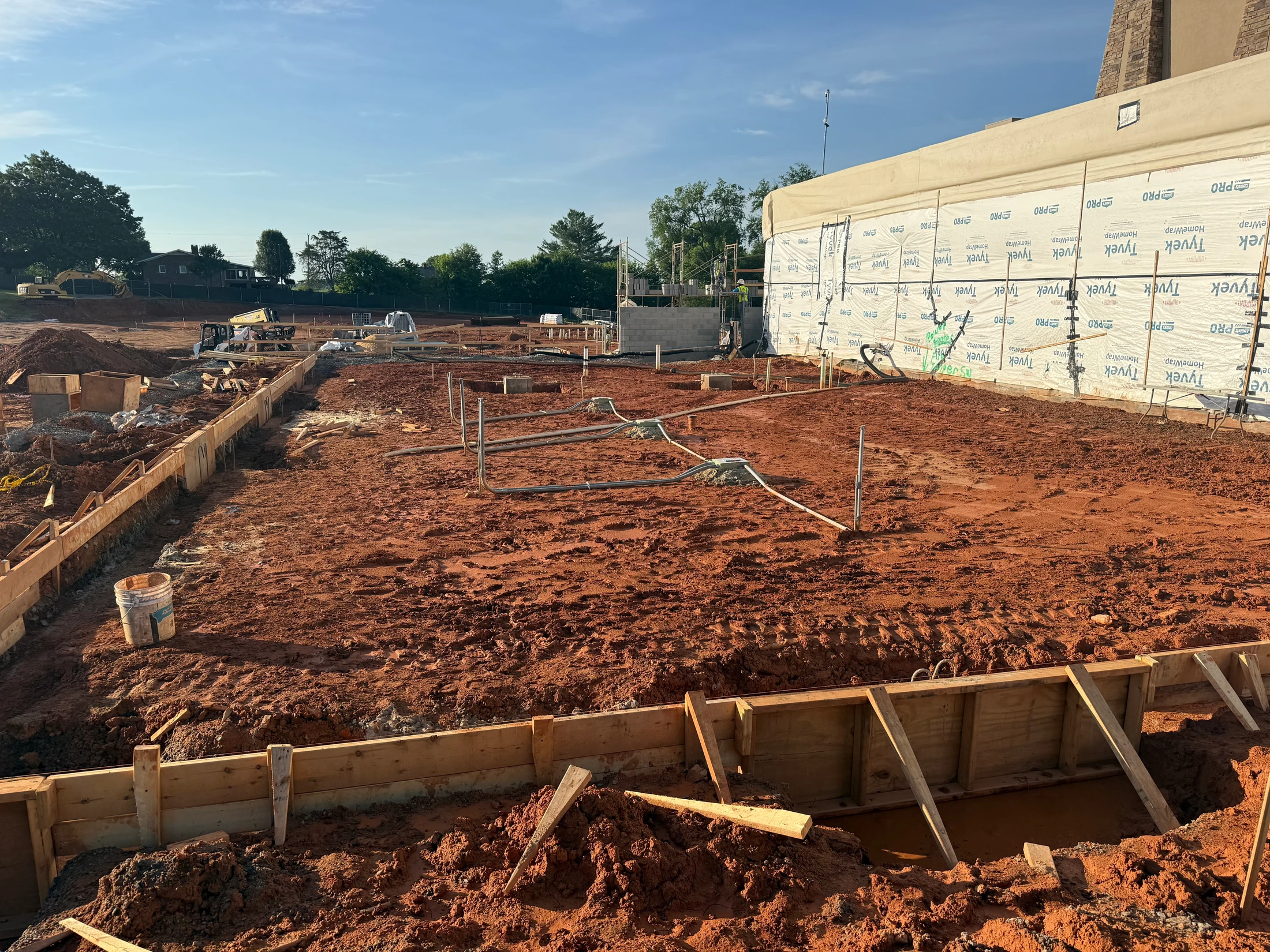 A construction site with red clay ground prepared for a foundation. A partially built structure is wrapped in white Tyvek, with wooden forms and pipes in place.