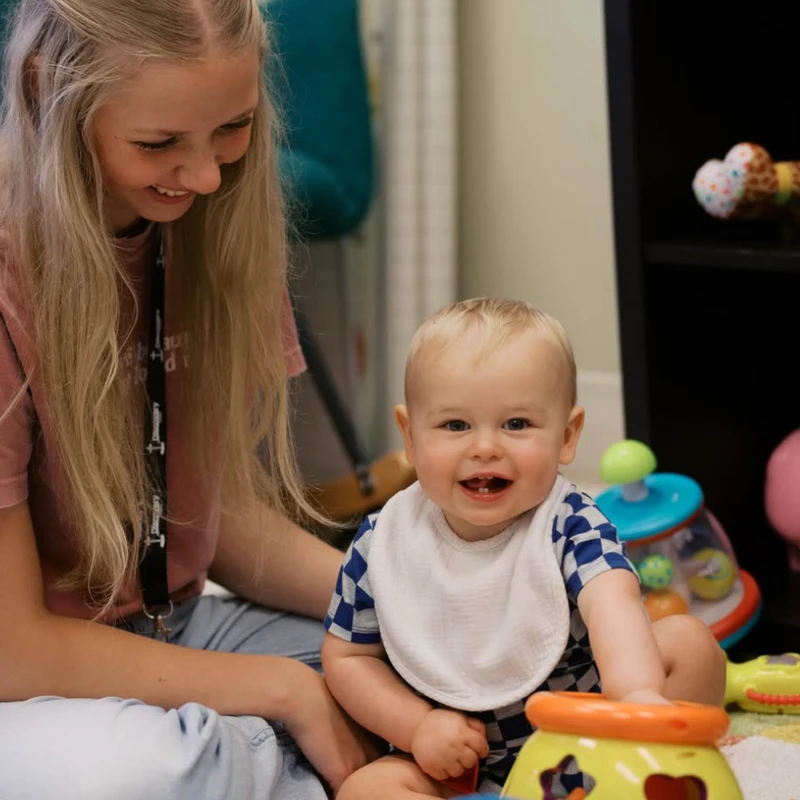 A happy baby with a white bib smiles at the camera while playing with toys on the floor next to a smiling blonde girl.