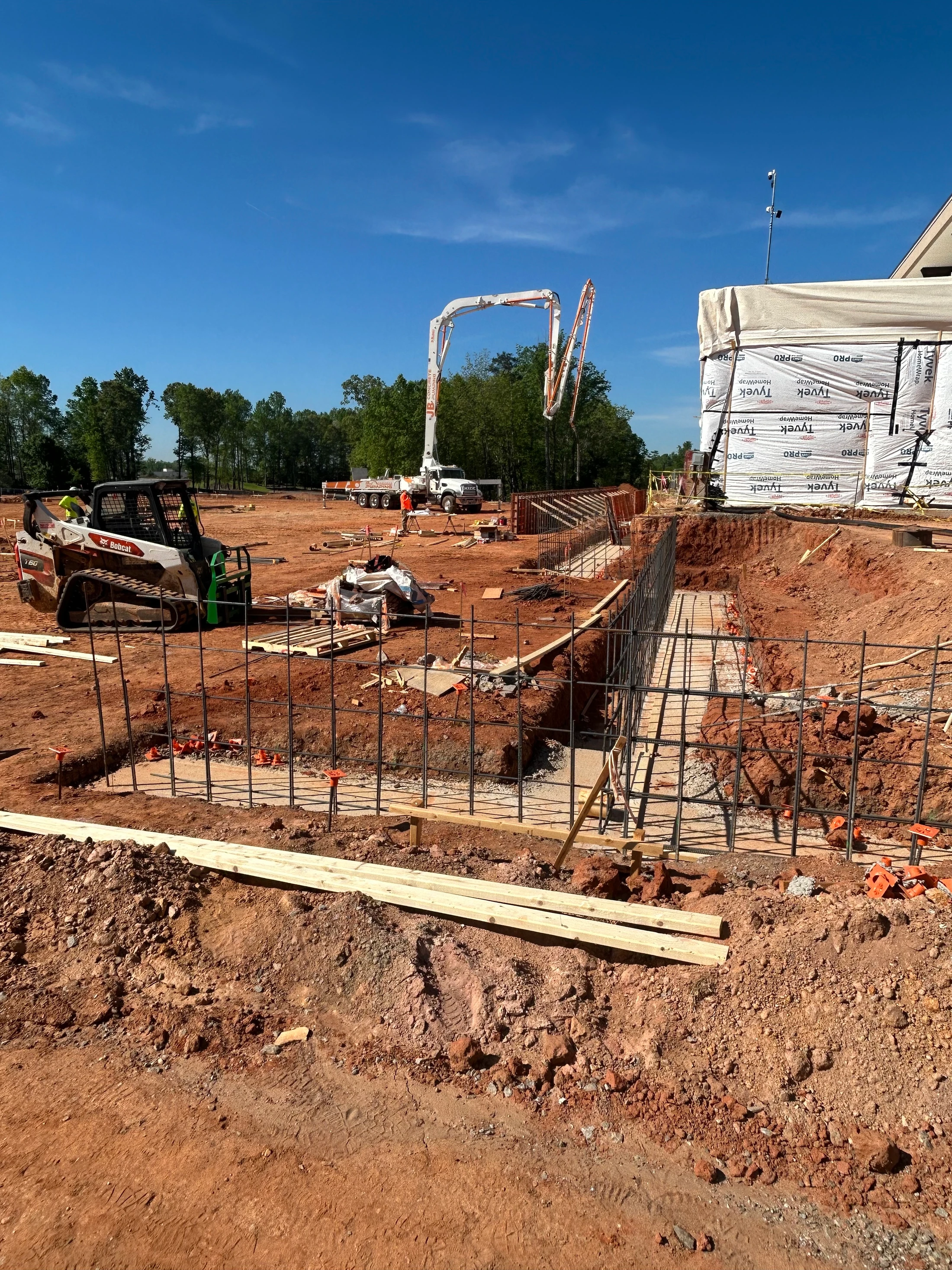 A large construction site on a sunny day. Rebar cages sit in a trench for a new foundation, with a Bobcat and a large concrete pump truck in the background.