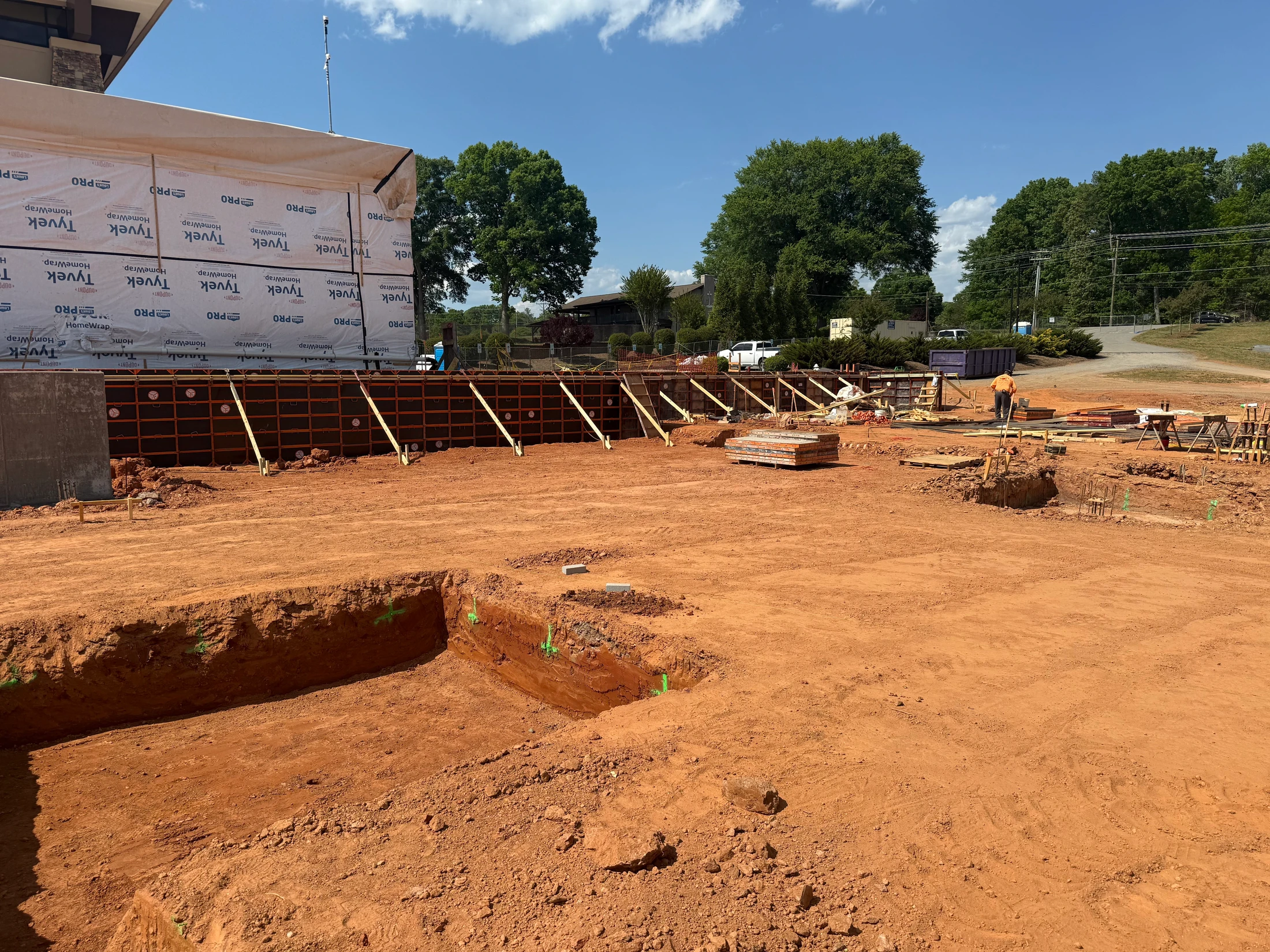 A wide shot of a construction site with red dirt ground. A building is being erected with concrete forms for a foundation wall set up on a sunny day.