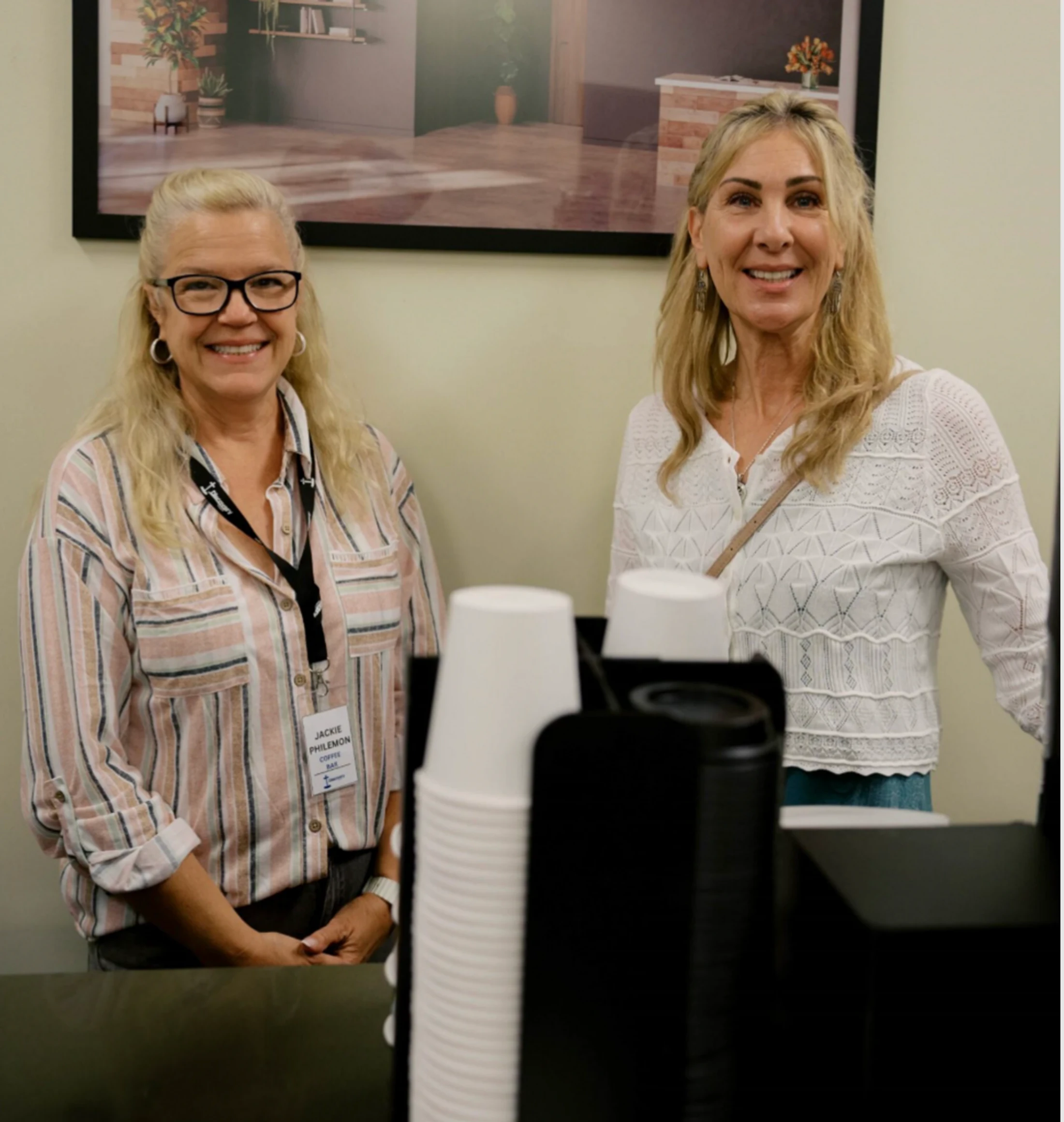 Two women smile for the camera while standing behind a counter with stacks of paper coffee cups. One wears glasses and a striped shirt with a name tag.