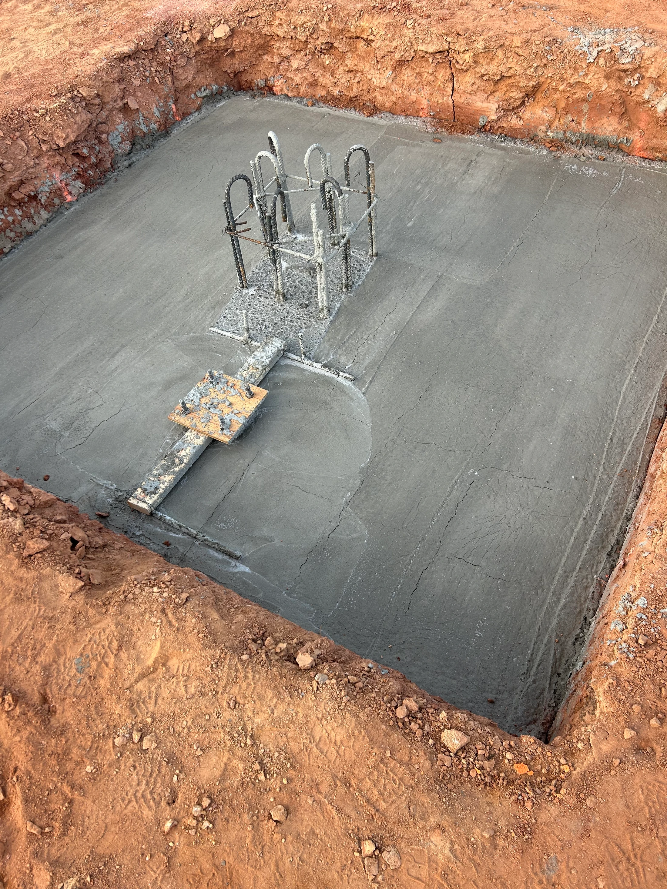 A square concrete foundation slab inside an earthen pit. A steel rebar cage for a future column protrudes from the center of the fresh concrete.