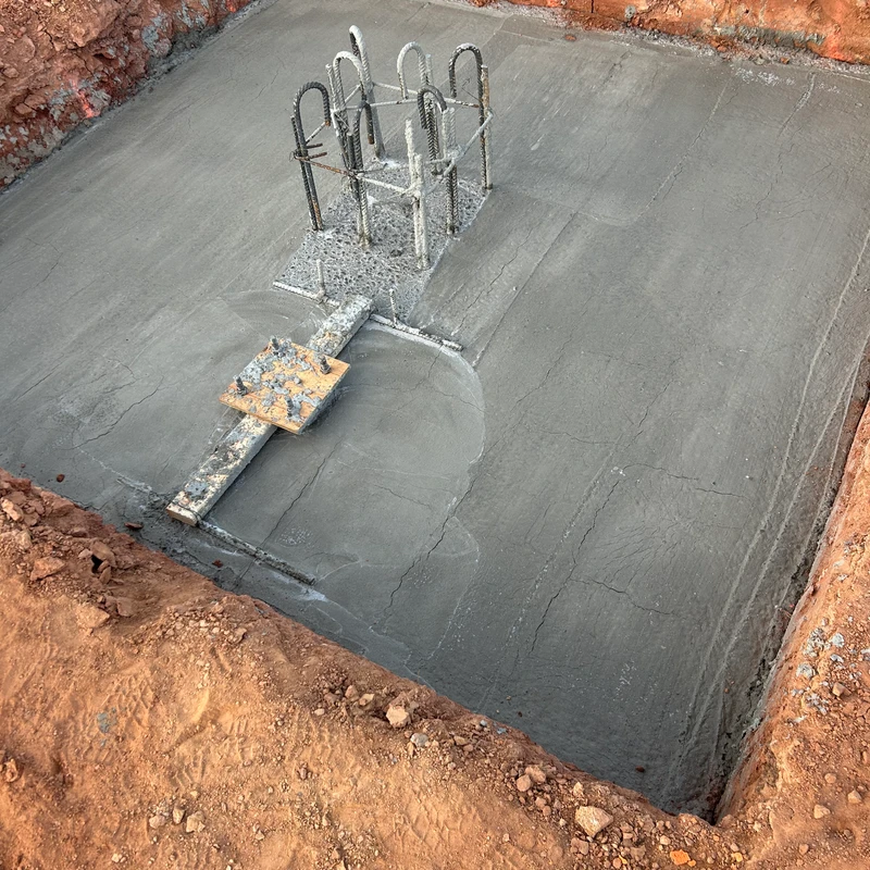 A square concrete foundation slab inside an earthen pit. A steel rebar cage for a future column protrudes from the center of the fresh concrete.