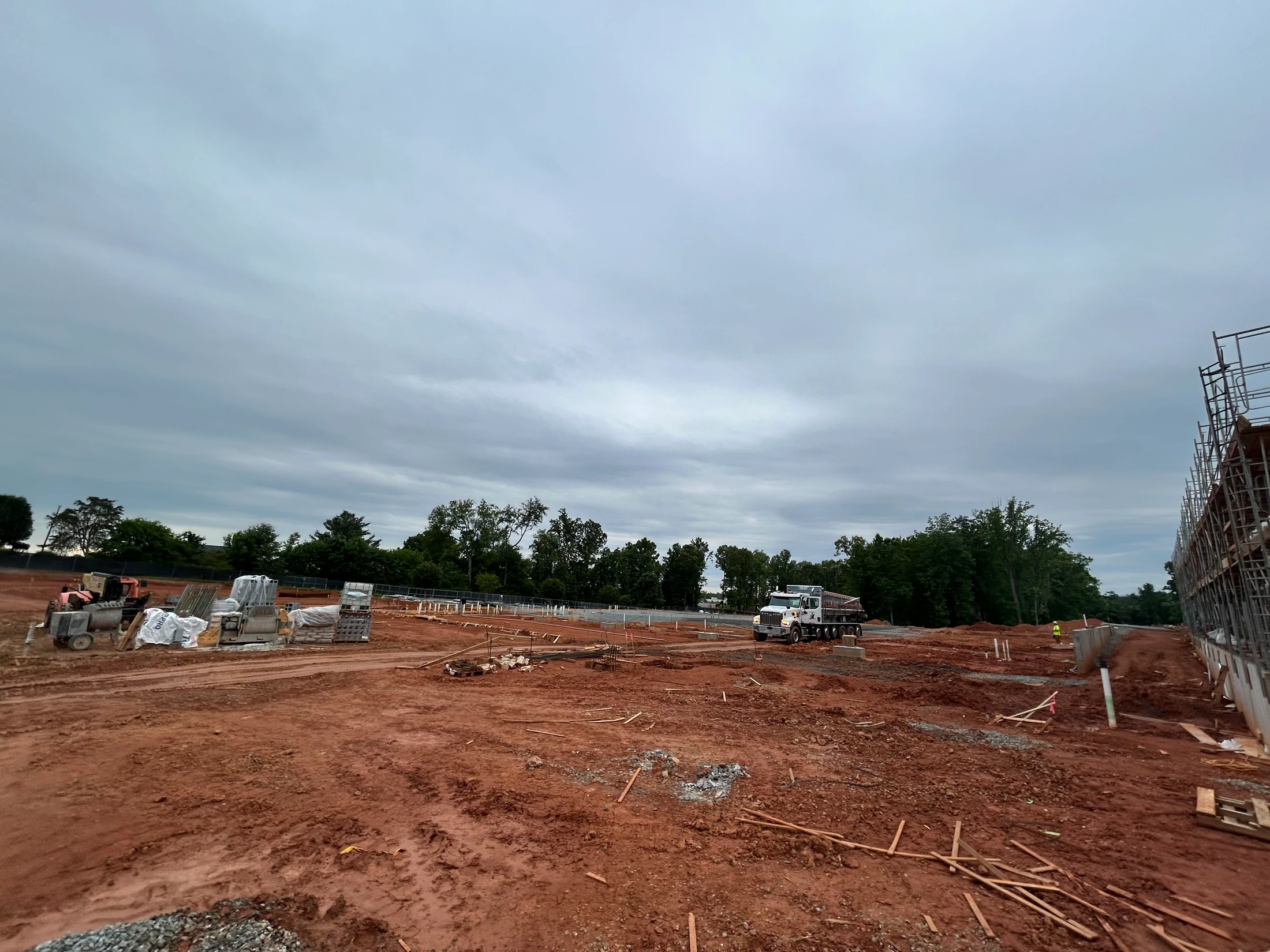 A wide view of a construction site with red, muddy ground under a grey, cloudy sky. Scaffolding, a large truck, and building materials are visible.