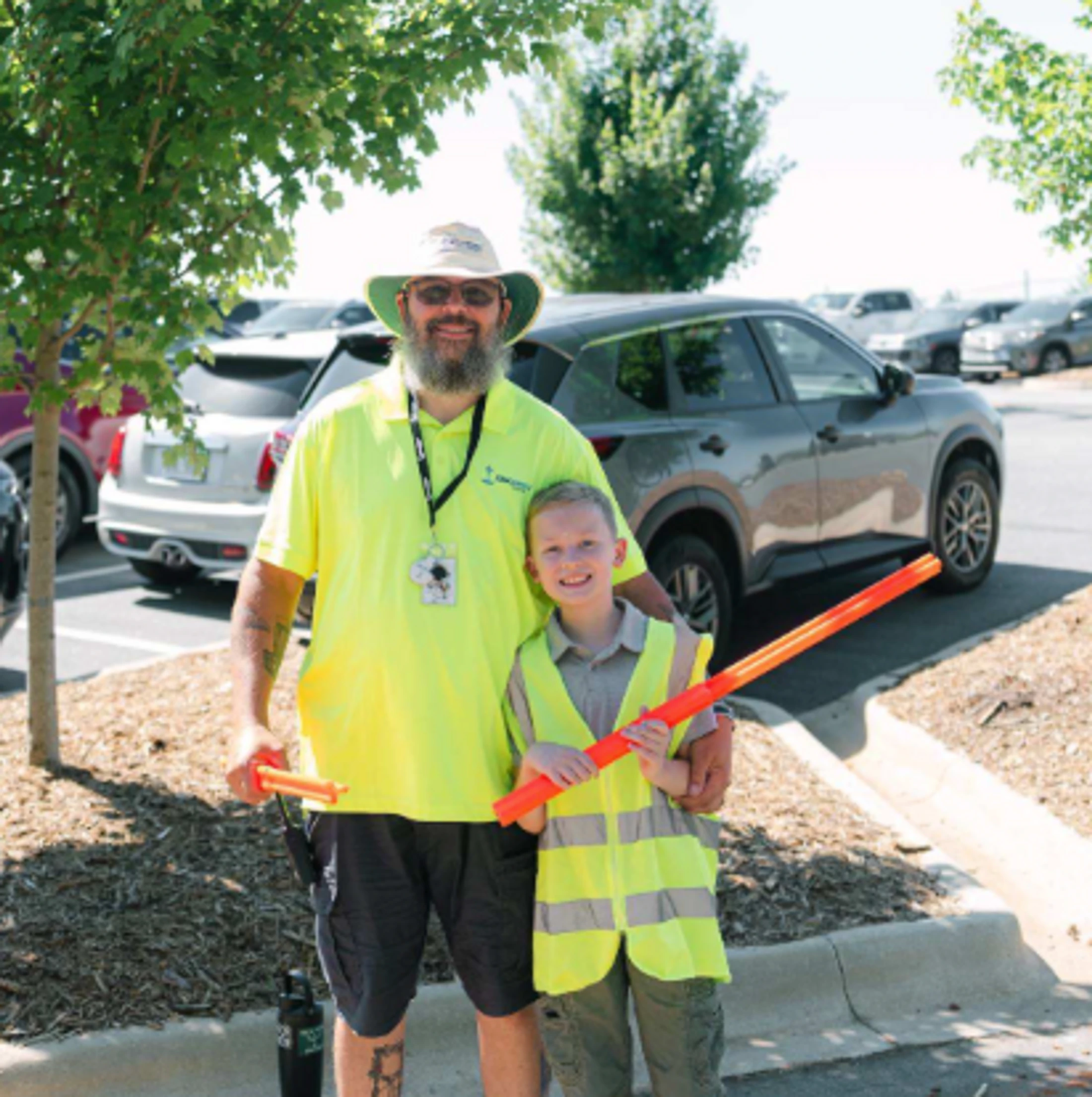 A man and a young boy in matching yellow safety vests smile while holding orange traffic wands in a sunny parking lot.