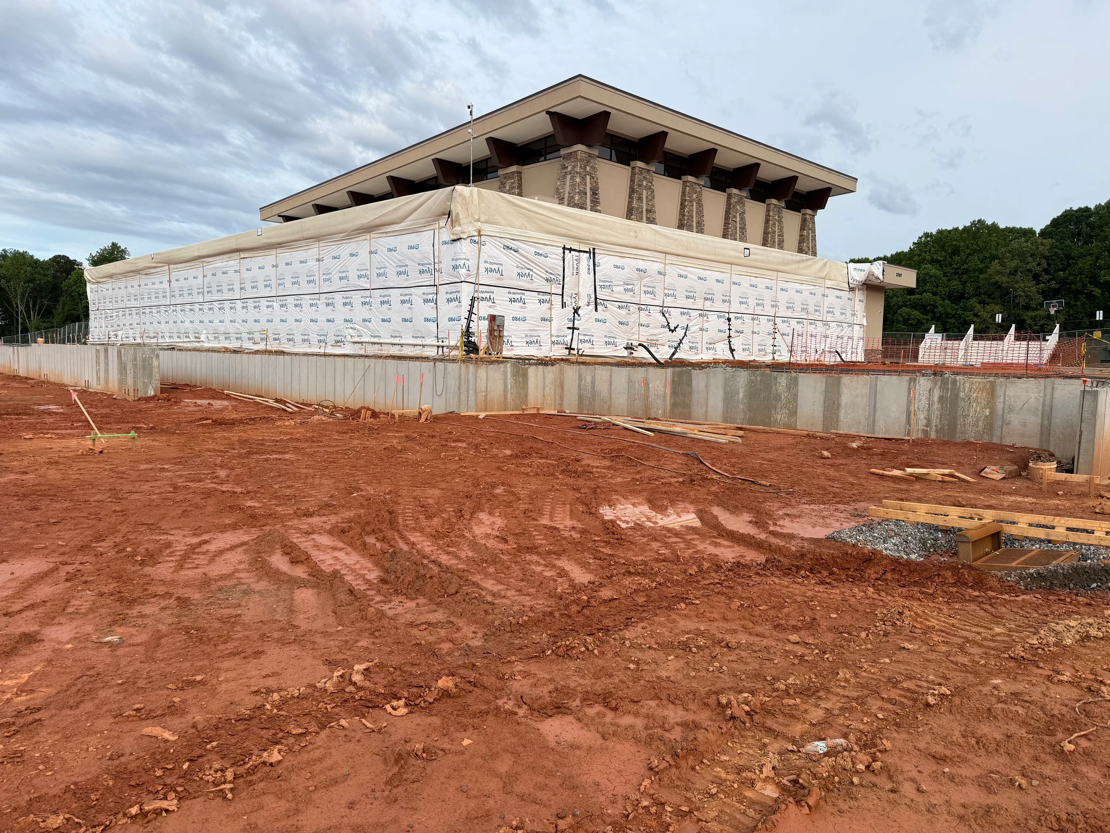 A construction site with wet red clay soil in the foreground and a large building with stone pillars under construction in the background, under a cloudy sky.