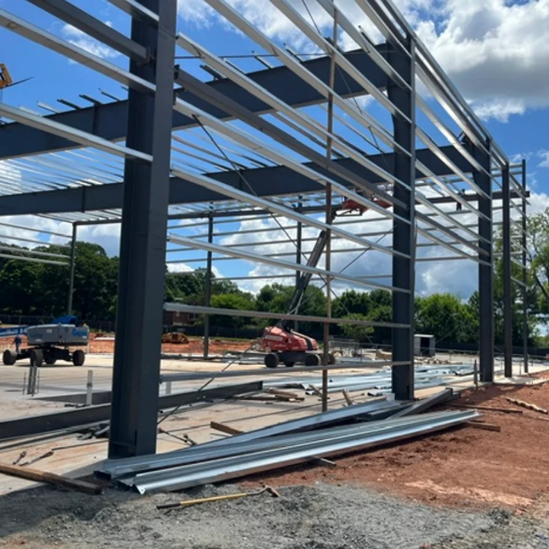 The steel frame of a large building under construction on a sunny day. Boom lifts and other equipment are visible on the dirt and concrete site.