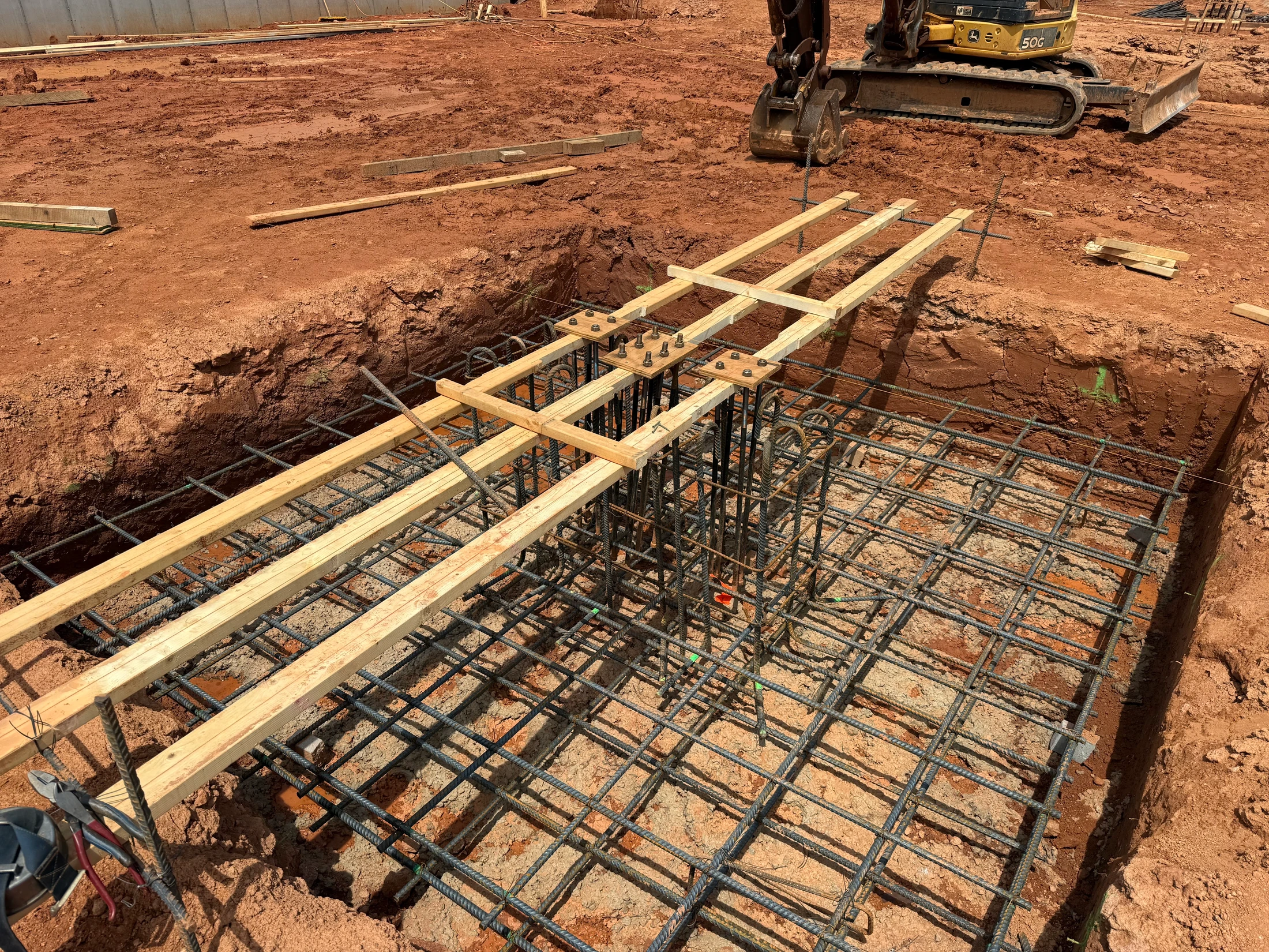 A construction site with red dirt, showing a trench with a steel rebar grid and column cage being prepared for a concrete foundation, with an excavator nearby.