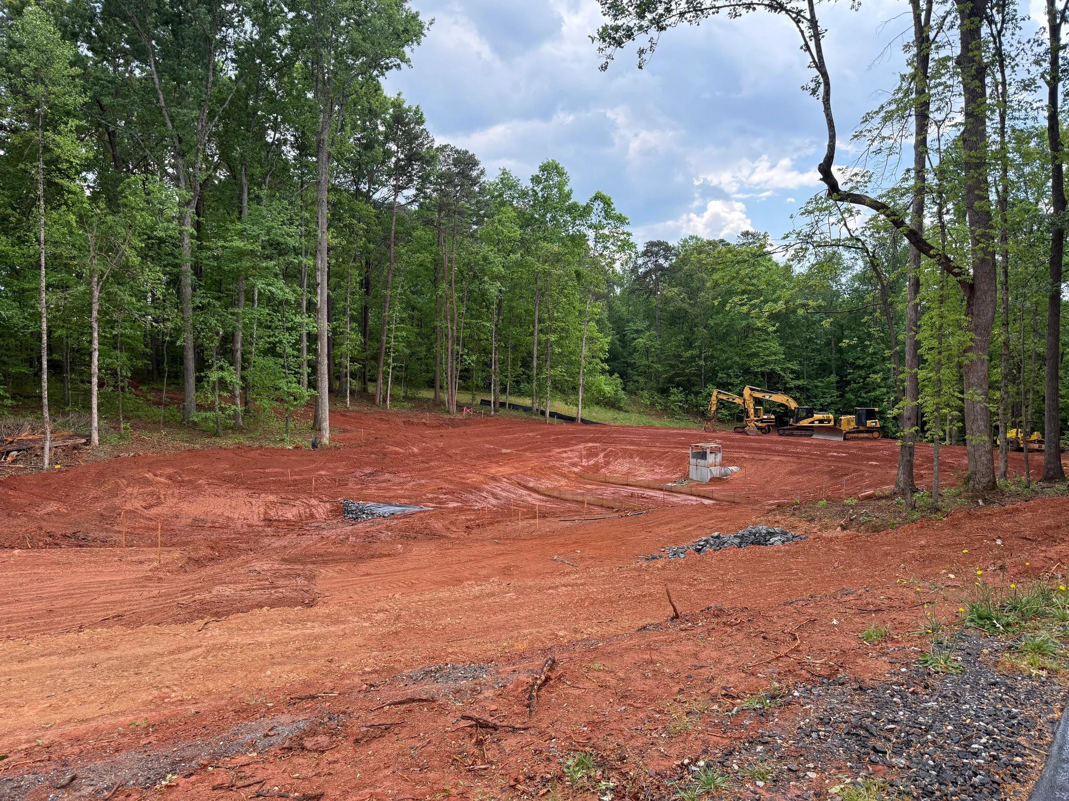 A large construction site with red clay soil graded and cleared. Heavy equipment sits at the edge of the surrounding forest under a cloudy sky.
