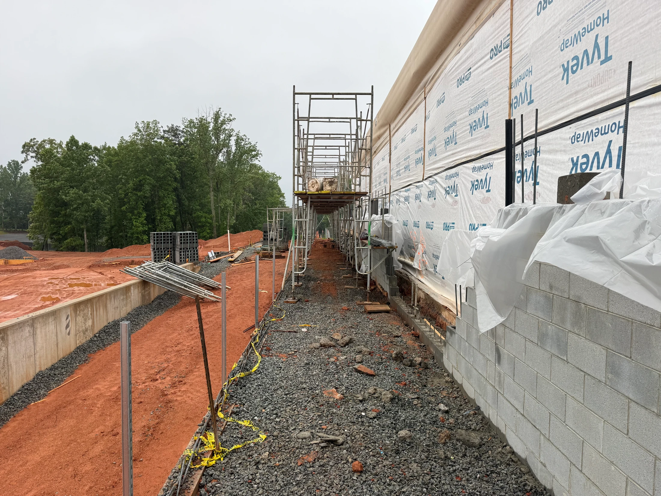 A view down the side of a building under construction, showing a partially built concrete block wall, white house wrap, and metal scaffolding on a gravel path.