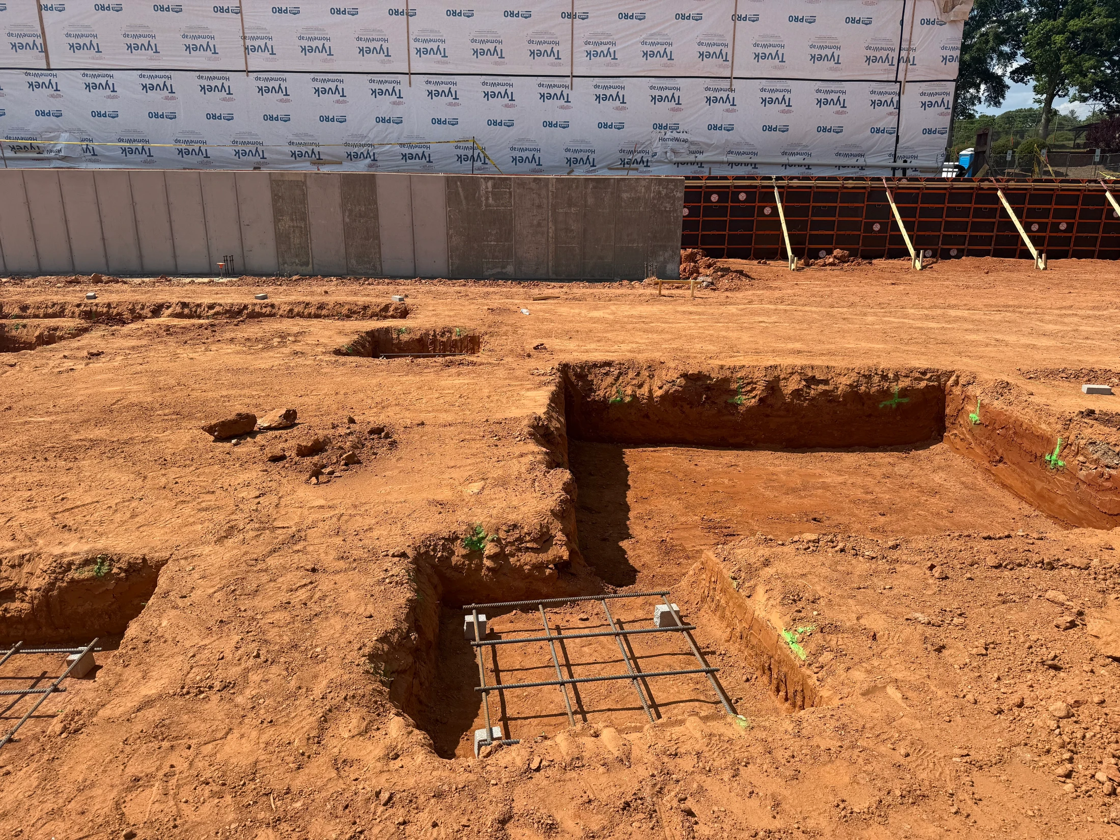 A construction site with excavated red earth. Rebar grids for concrete footings sit in dug-out squares. A concrete wall with Tyvek wrap is in the background.