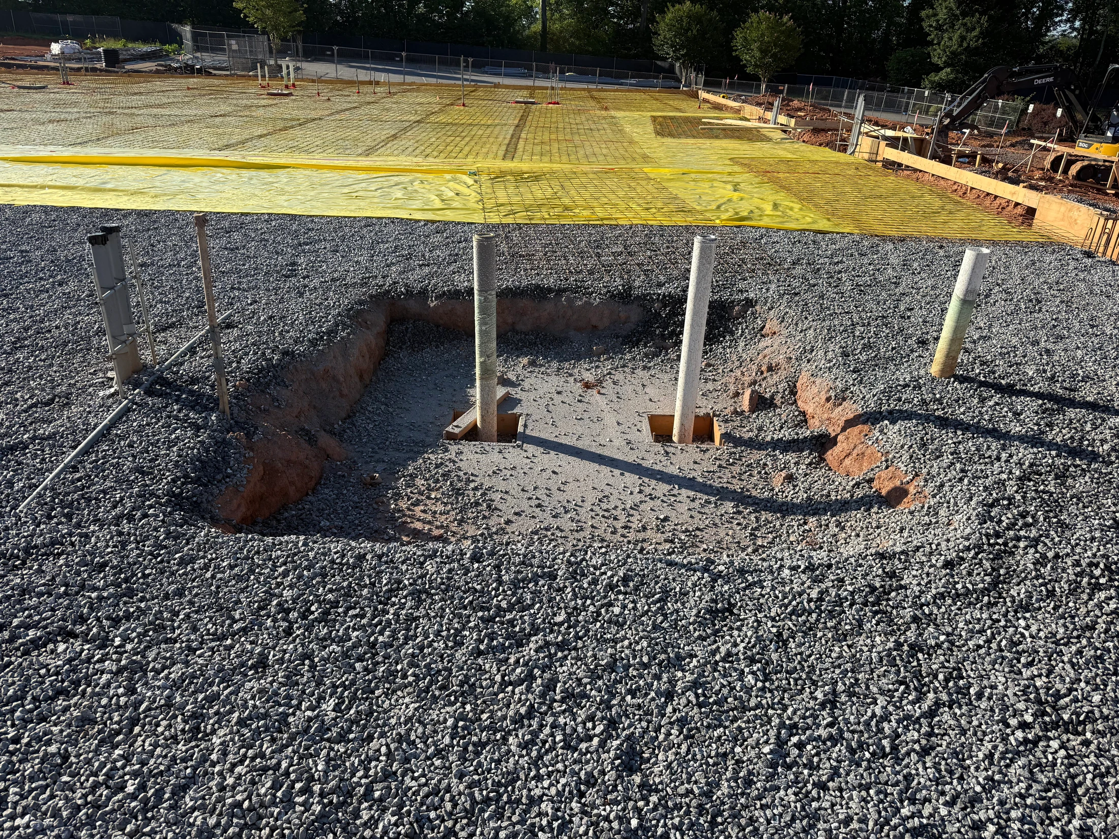 A construction site with a gravel bed. A pit in the foreground shows concrete footings, and in the background, a rebar grid lies on a yellow vapor barrier.