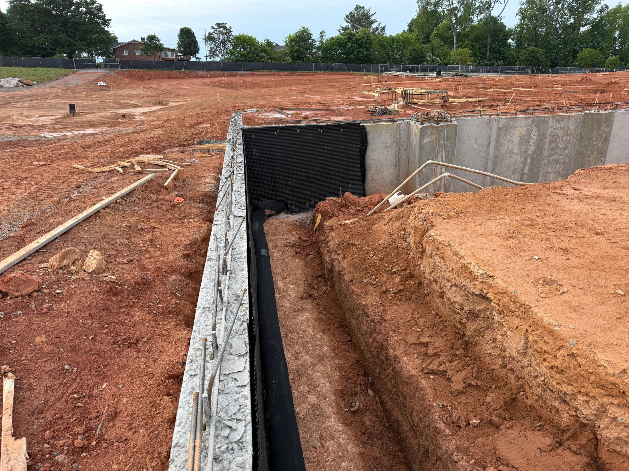 A construction site with an unfinished concrete foundation. A trench runs alongside a wall with black waterproofing and exposed rebar on red clay soil.