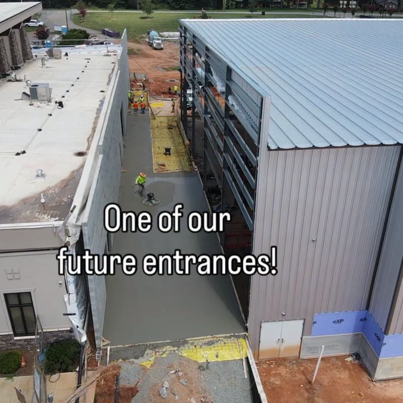 An aerial view of a construction site where workers are pouring and finishing a concrete walkway between two buildings for a future entrance.