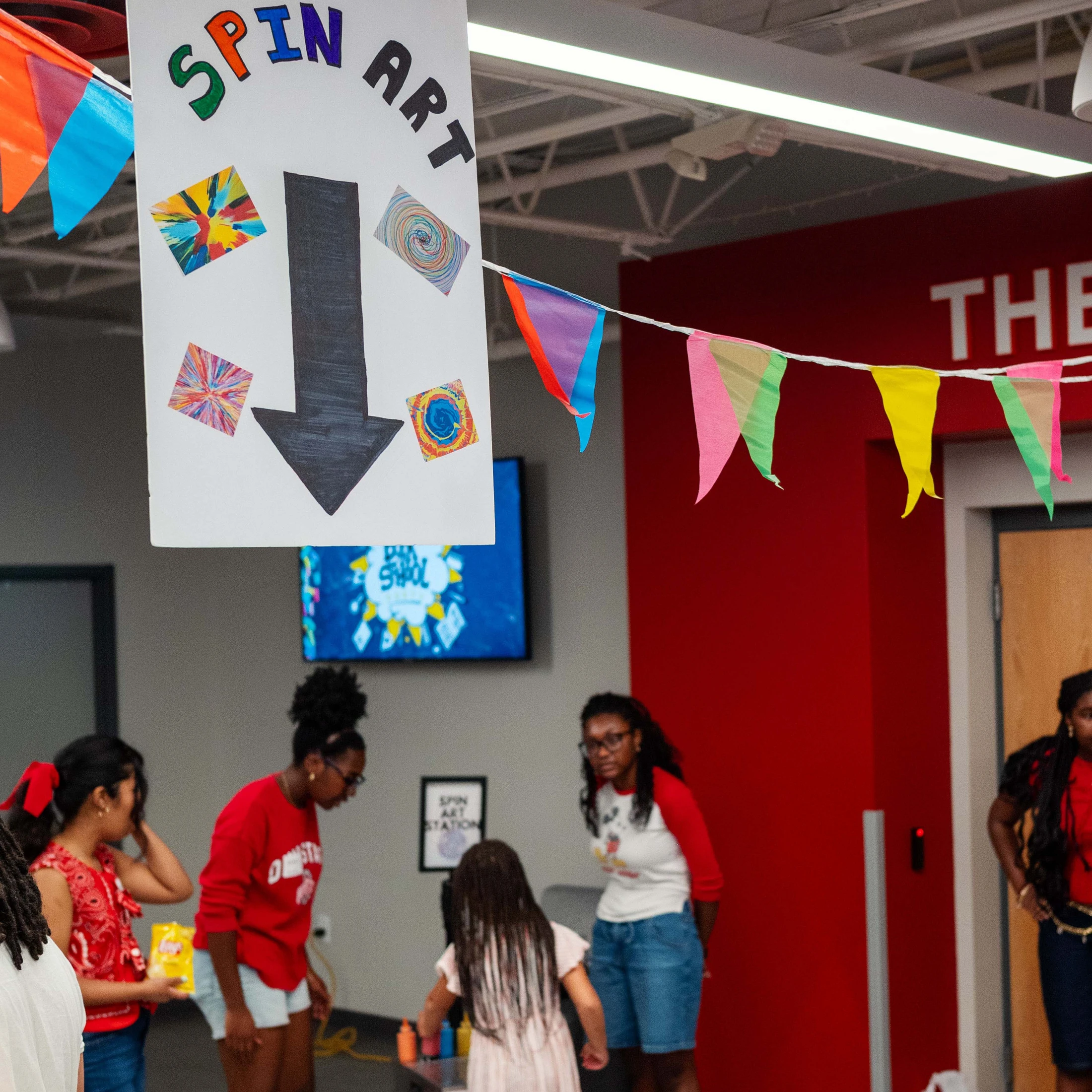A handmade 'Spin Art' sign with a large arrow hangs from the ceiling, pointing down towards a group of young people at an activity station.