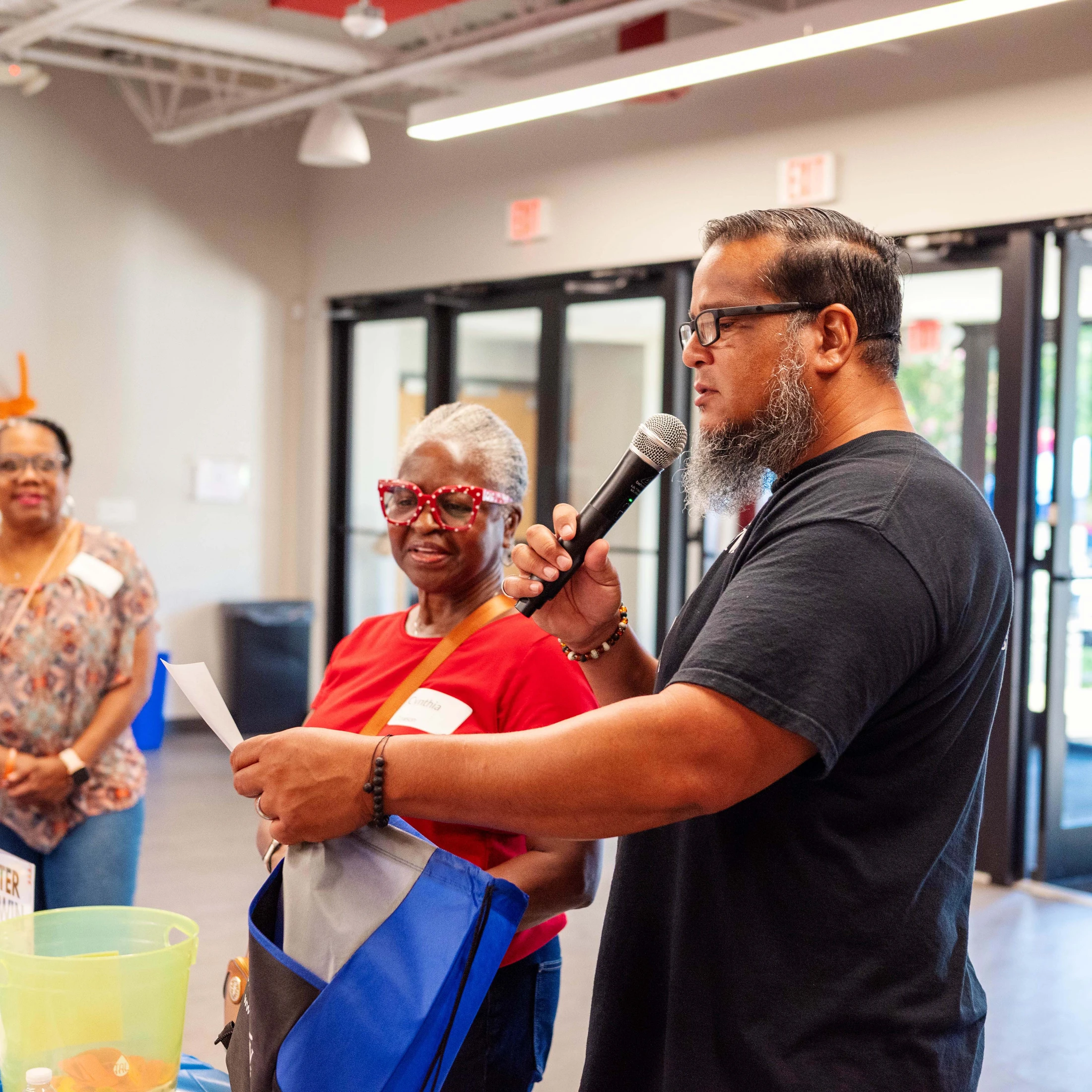 A man with a beard and glasses speaks into a microphone at an indoor gathering, as two women stand behind him listening attentively.