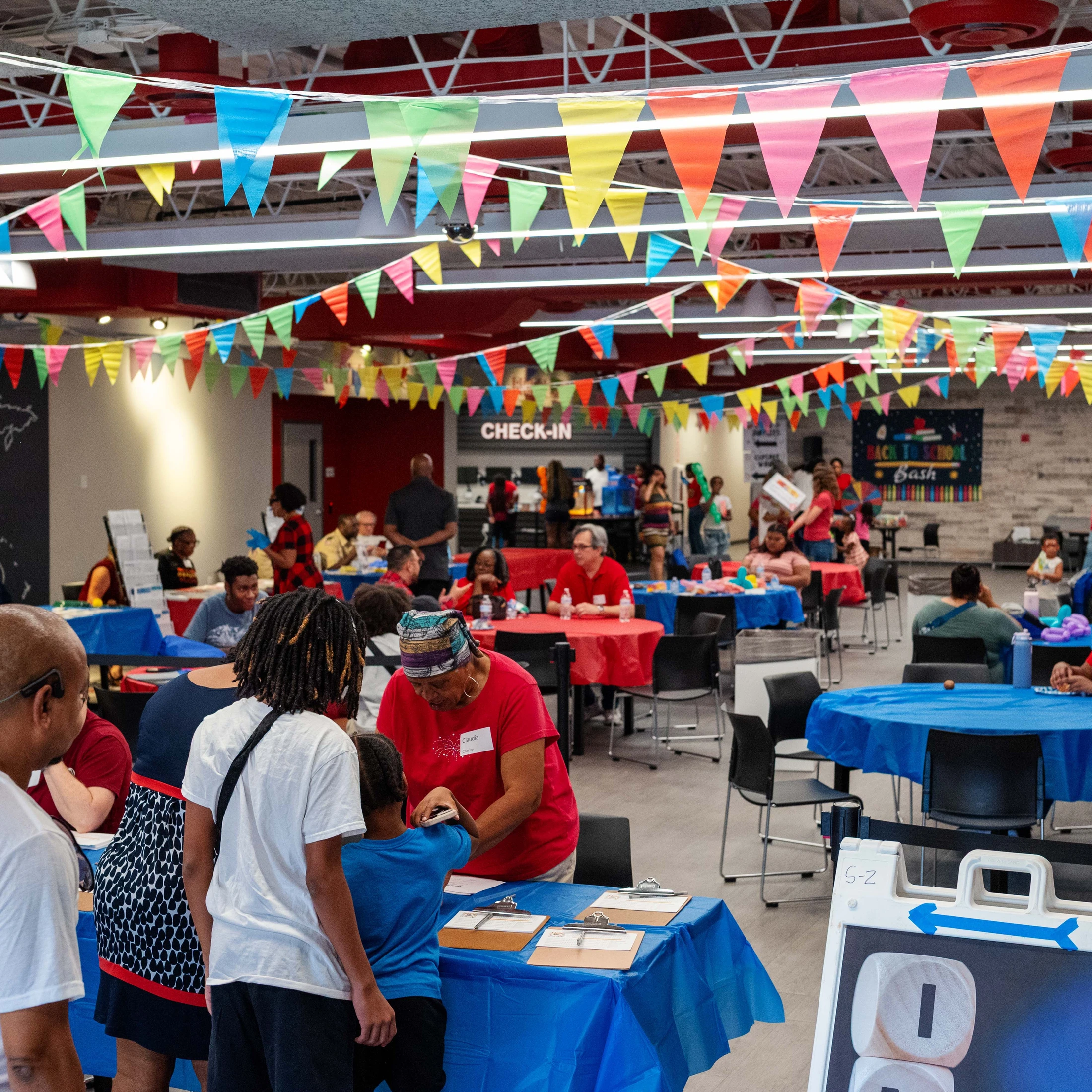 A large room full of families and volunteers at a back-to-school event, with colorful banners hanging from the ceiling and registration tables in the front.