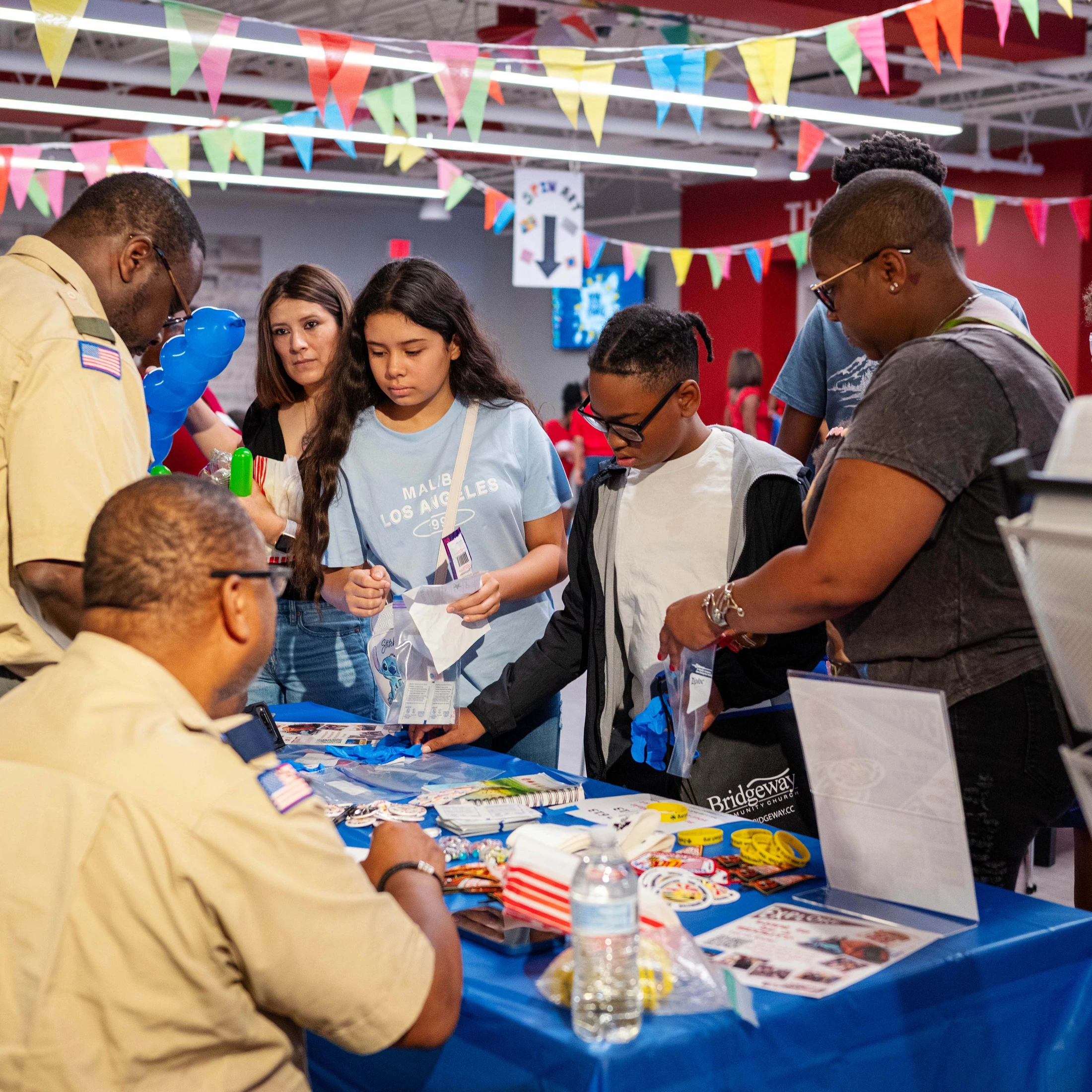 A family with two children interact with two men in scout uniforms at a booth during an indoor event with colorful banners overhead.