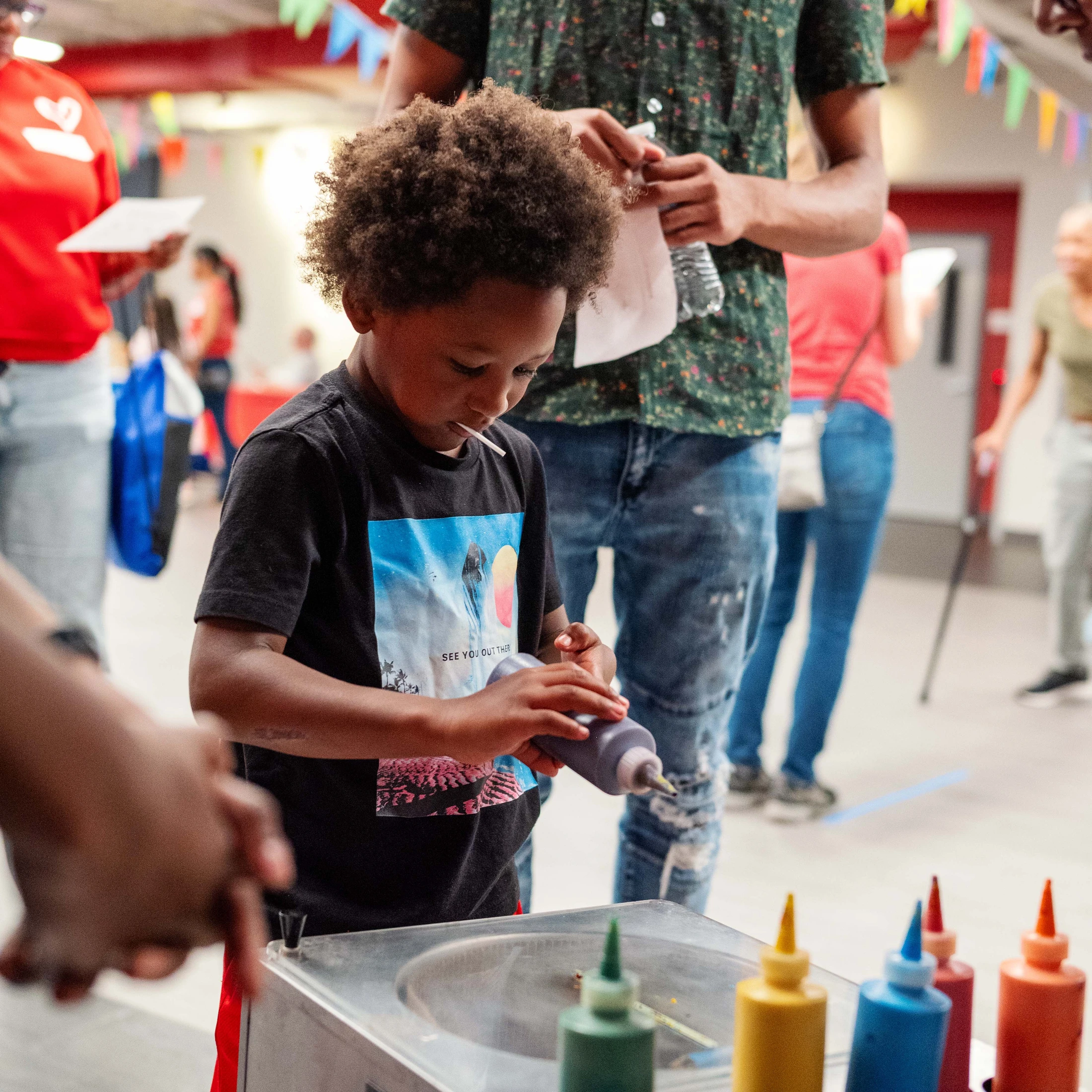 A young boy with curly hair and a lollipop in his mouth concentrates on squeezing grey paint from a bottle onto a spin art machine at a crowded event.