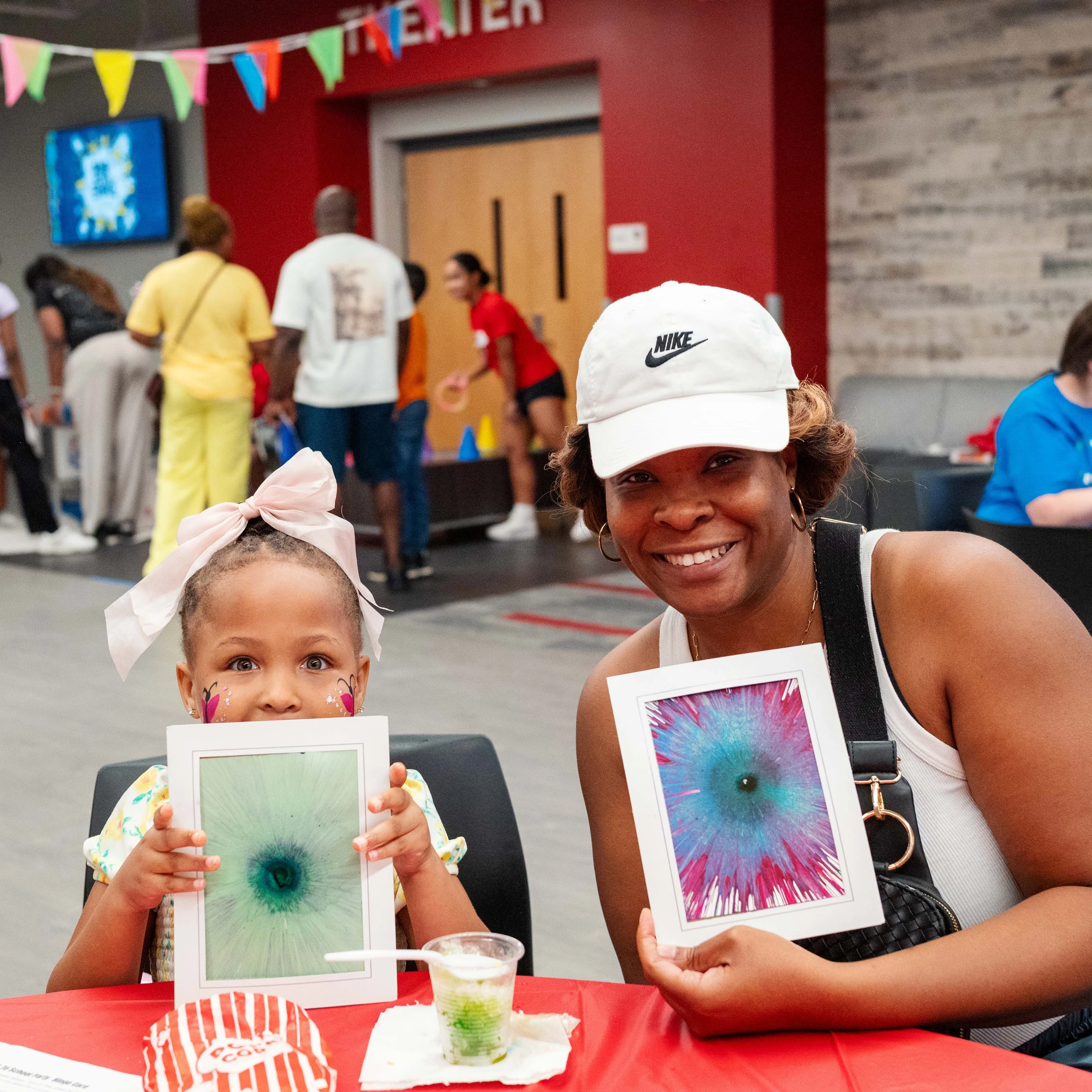 A smiling woman and a young girl with butterfly face paint sit at a red table, happily holding up their colorful, framed spin art creations at an event.