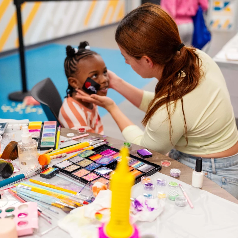 A woman gently paints a design on a young girl's cheek. In front of them, a table is covered with colorful face painting supplies like palettes and glitter.