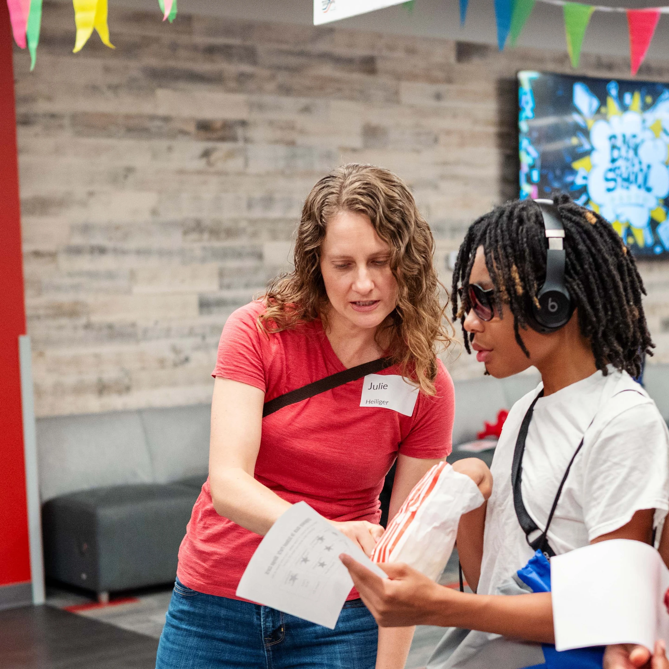 A woman in a red shirt with a nametag points to a paper while talking to a young person with dreadlocks and headphones at a back-to-school event.