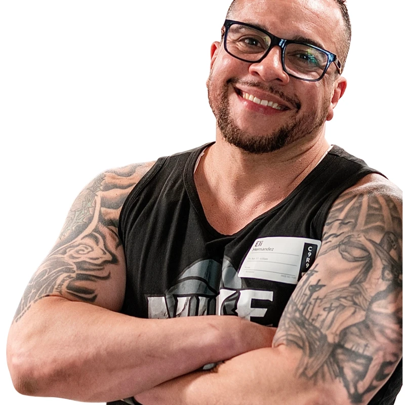 A muscular man with glasses, a beard, and tattoos on his arms smiles at the camera. He is wearing a black tank top and has his arms crossed.