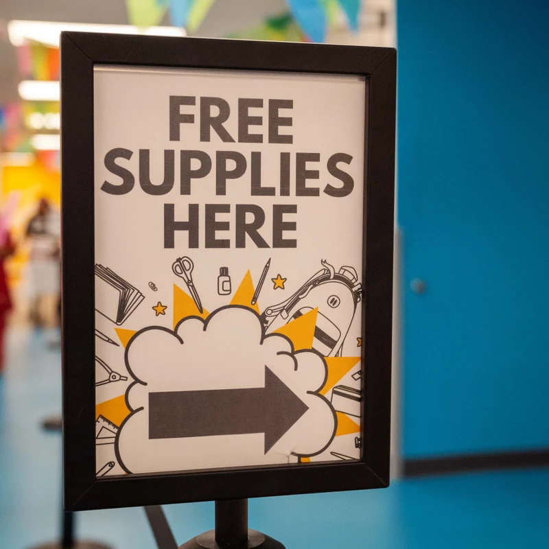 A black-framed sign on a stand reads 'FREE SUPPLIES HERE' with a large arrow pointing right, set in a colorful hallway with banners.