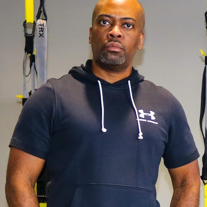 A bald Black man with a goatee wearing a dark blue short-sleeved Under Armour hoodie, posing in front of yellow TRX suspension trainers in a gym.
