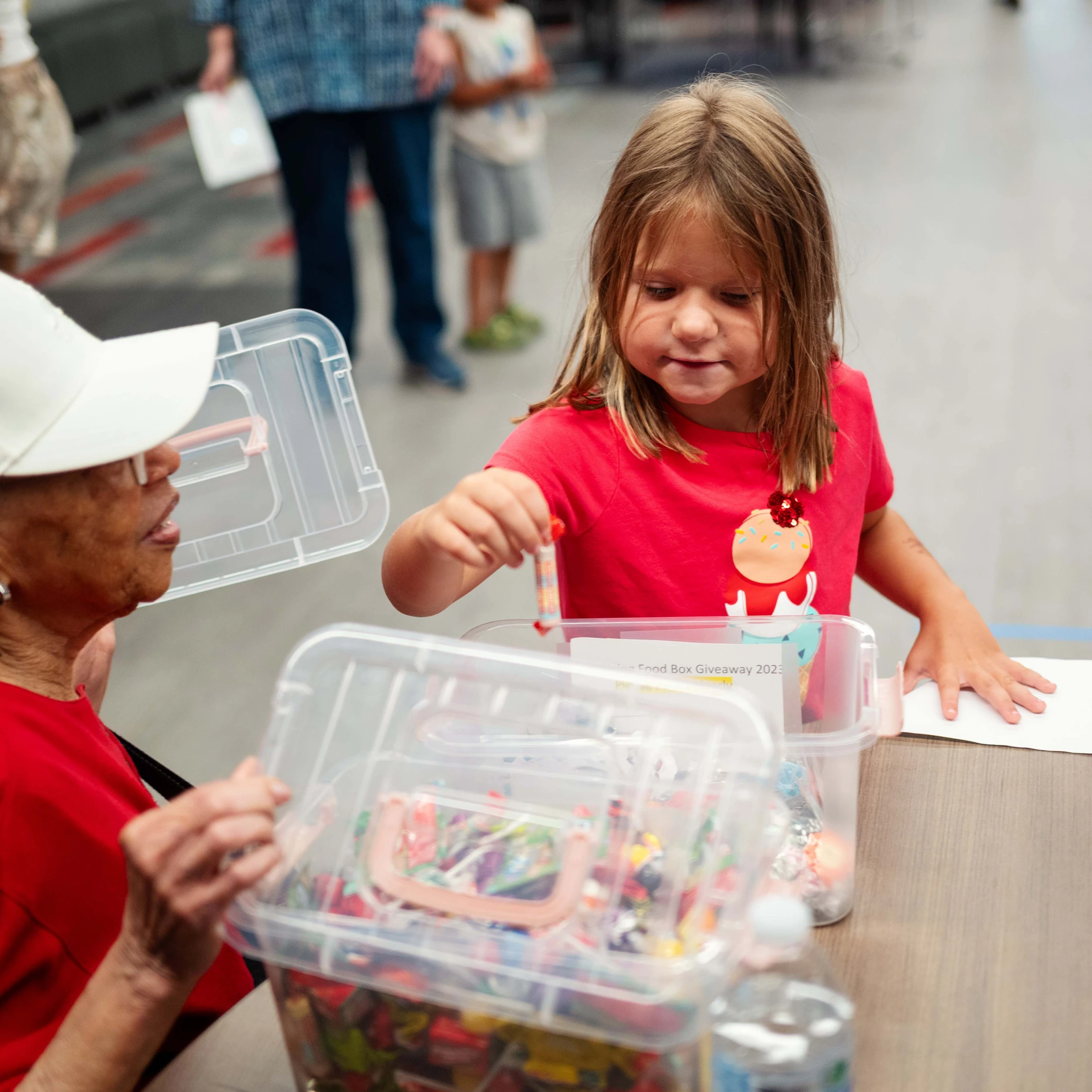 A young girl in a red shirt selects a piece of candy from a plastic tub held by an older woman wearing a white hat at a community giveaway.