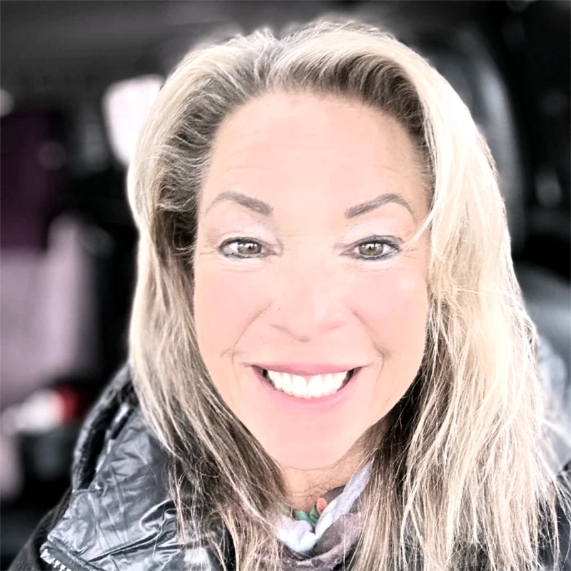 A close-up, bright selfie of a smiling woman with blonde hair. She is wearing a black puffy jacket and a scarf, and the background is blurred.