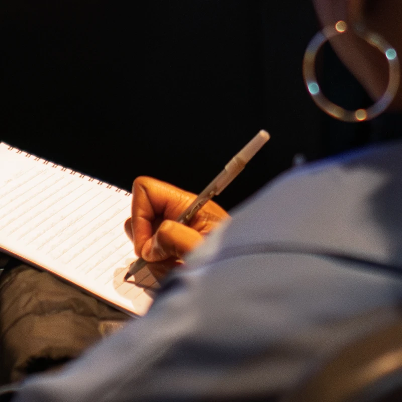 An over-the-shoulder, close-up shot of a woman's hand writing with a pen in a lined, spiral notebook. Her large hoop earring is visible in the background.
