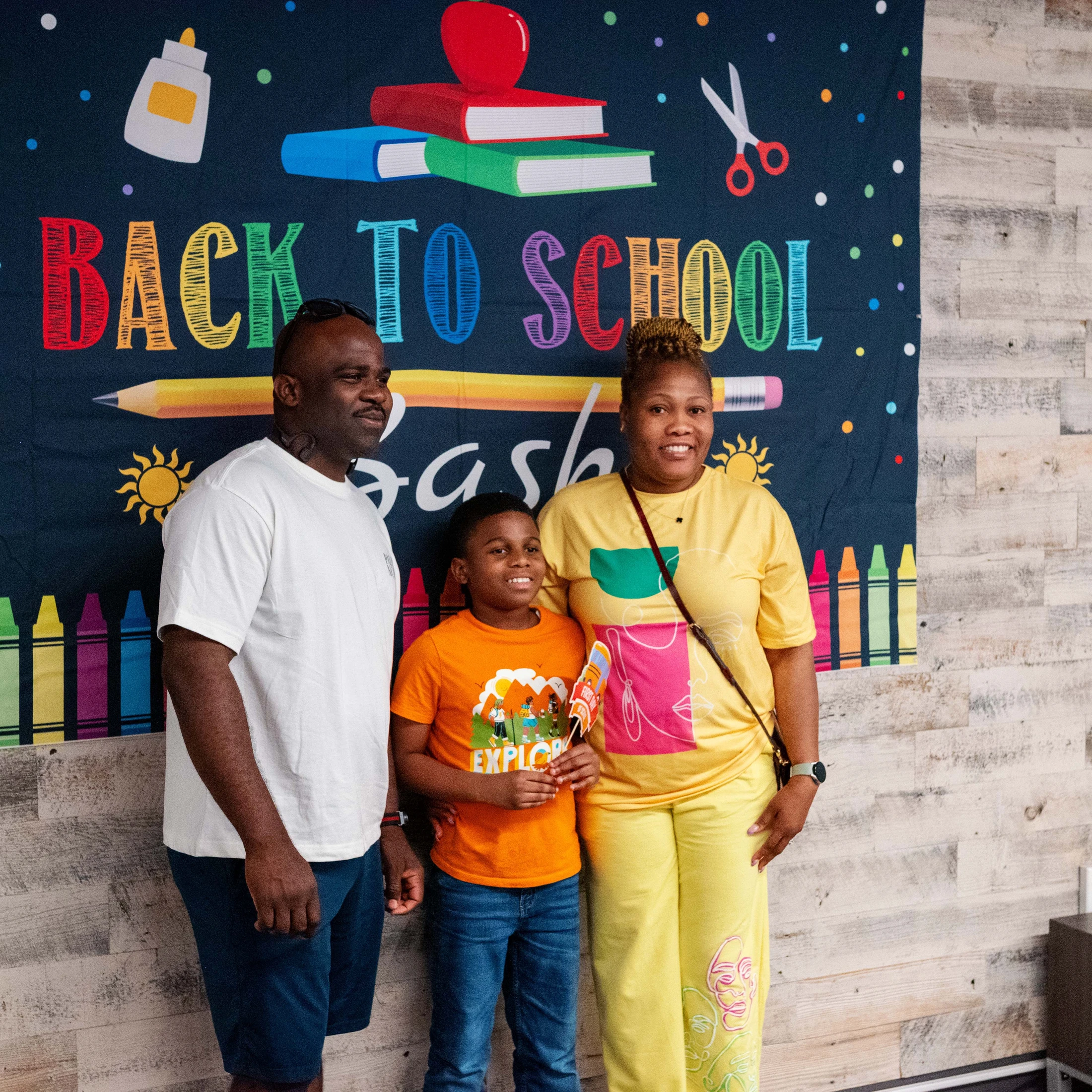 A smiling Black family with their son posing for a photo in front of a colorful 'Back to School Bash' banner with illustrations of school supplies.