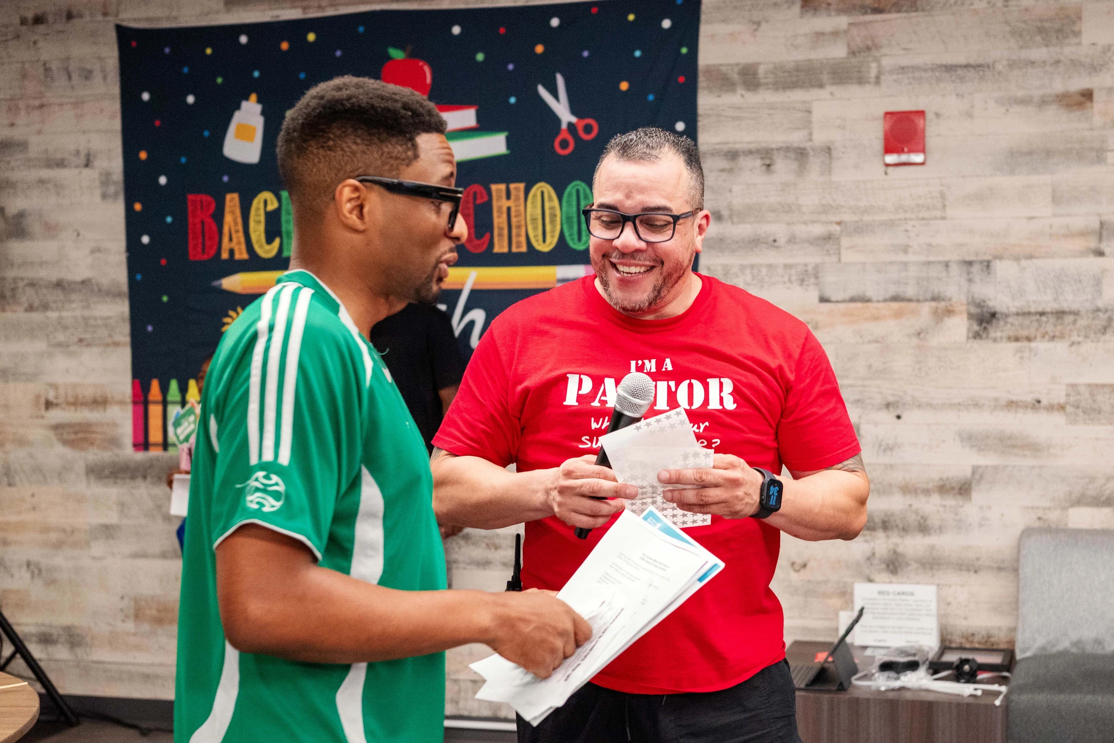 A smiling man in a red 'Pastor' shirt holds a microphone and talks with another man in a green shirt, in front of a 'Back to School' banner.