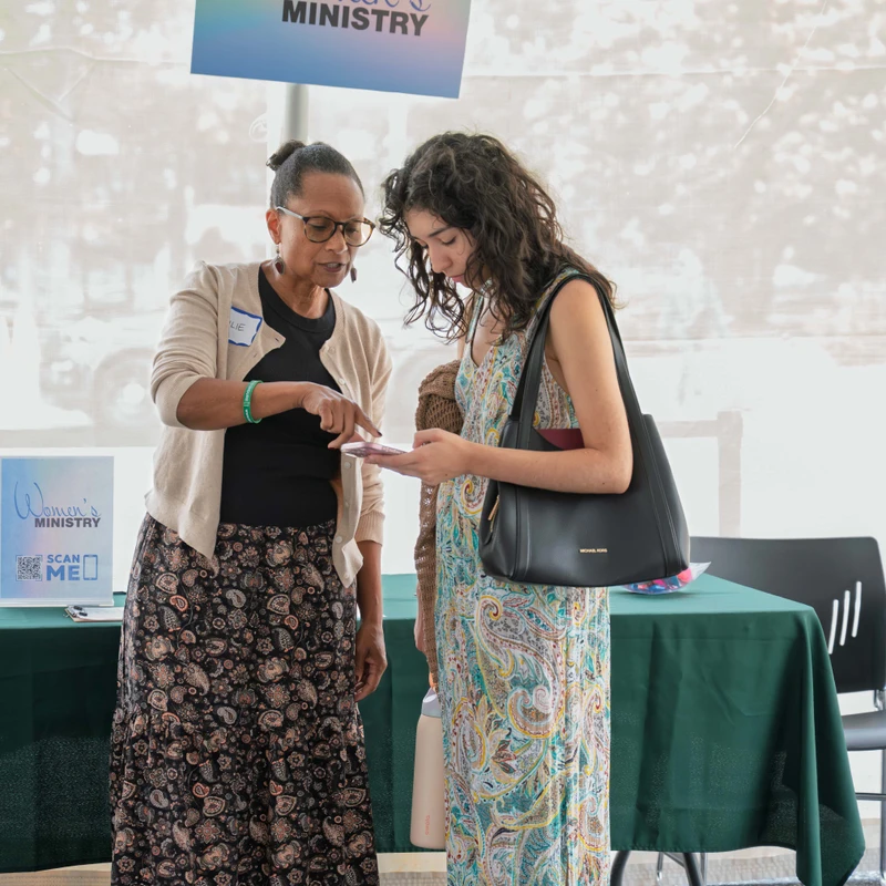 An older woman with glasses points to a phone held by a younger woman as they talk under a tent at a Women's Ministry event booth.