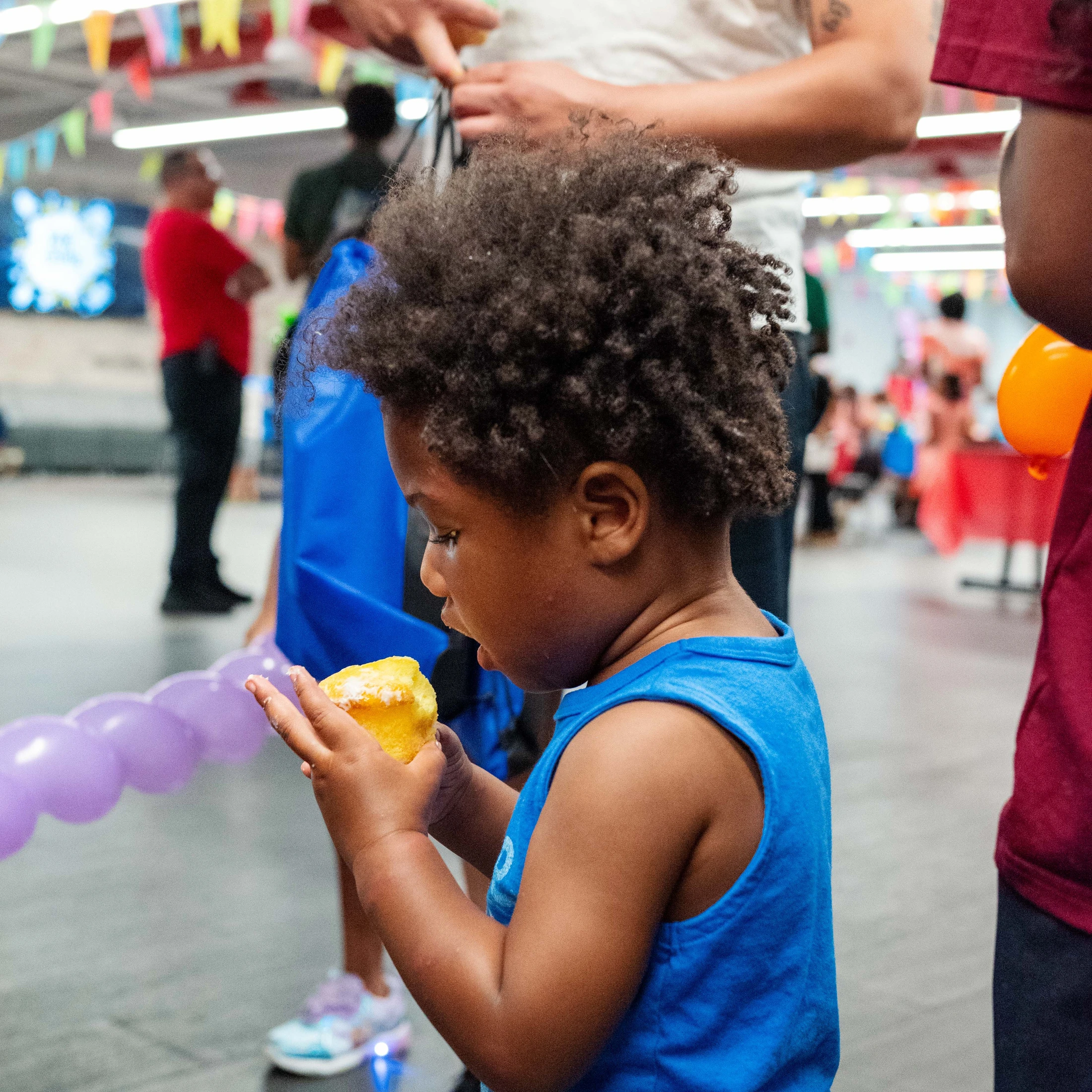A young Black child in a blue tank top stands indoors at a party, holding a small yellow cupcake with both hands and taking a bite.
